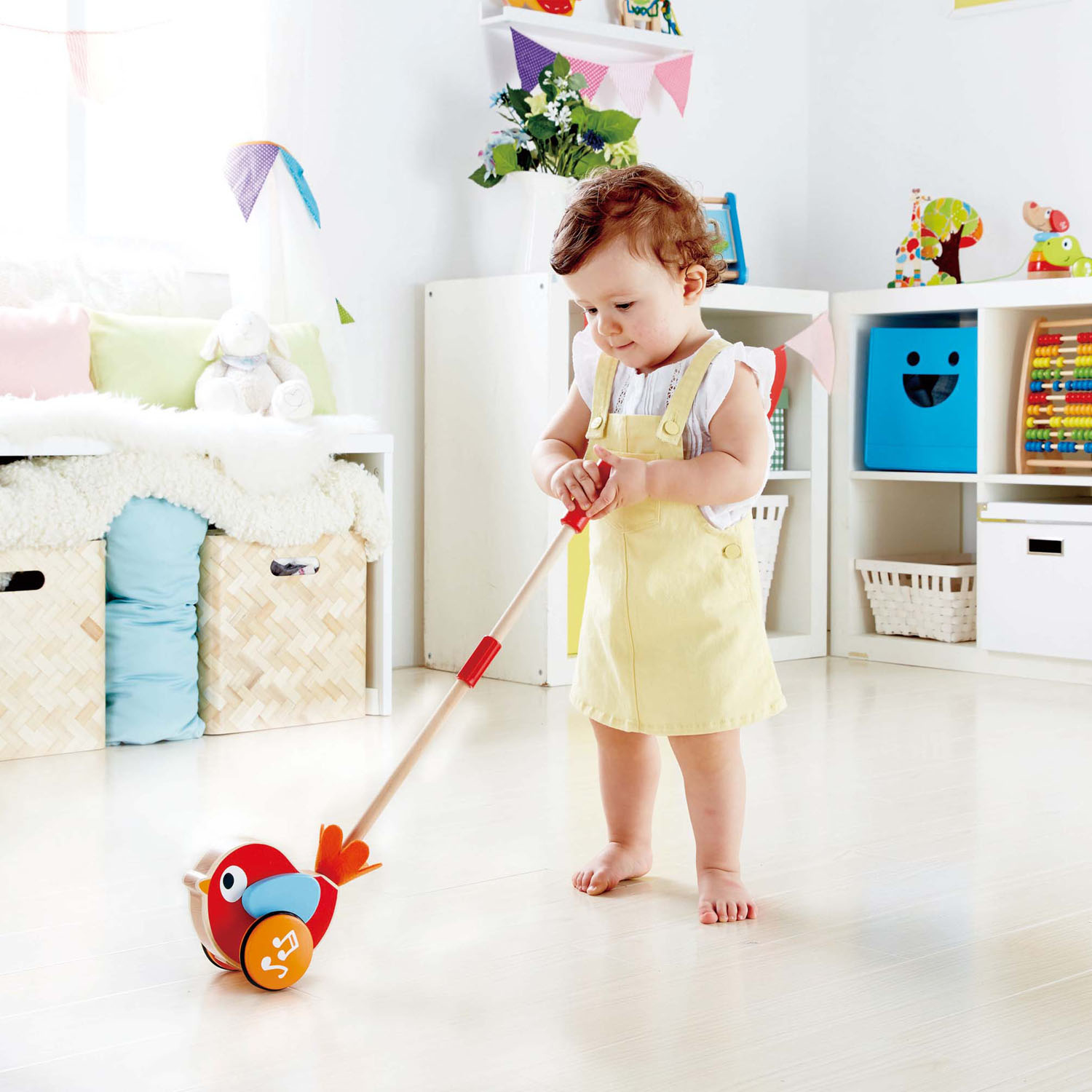 Niemowlę pcha czerwoną i pomarańczową zabawkę w kształcie ptaka na kółkach. Stoją w pokoju.