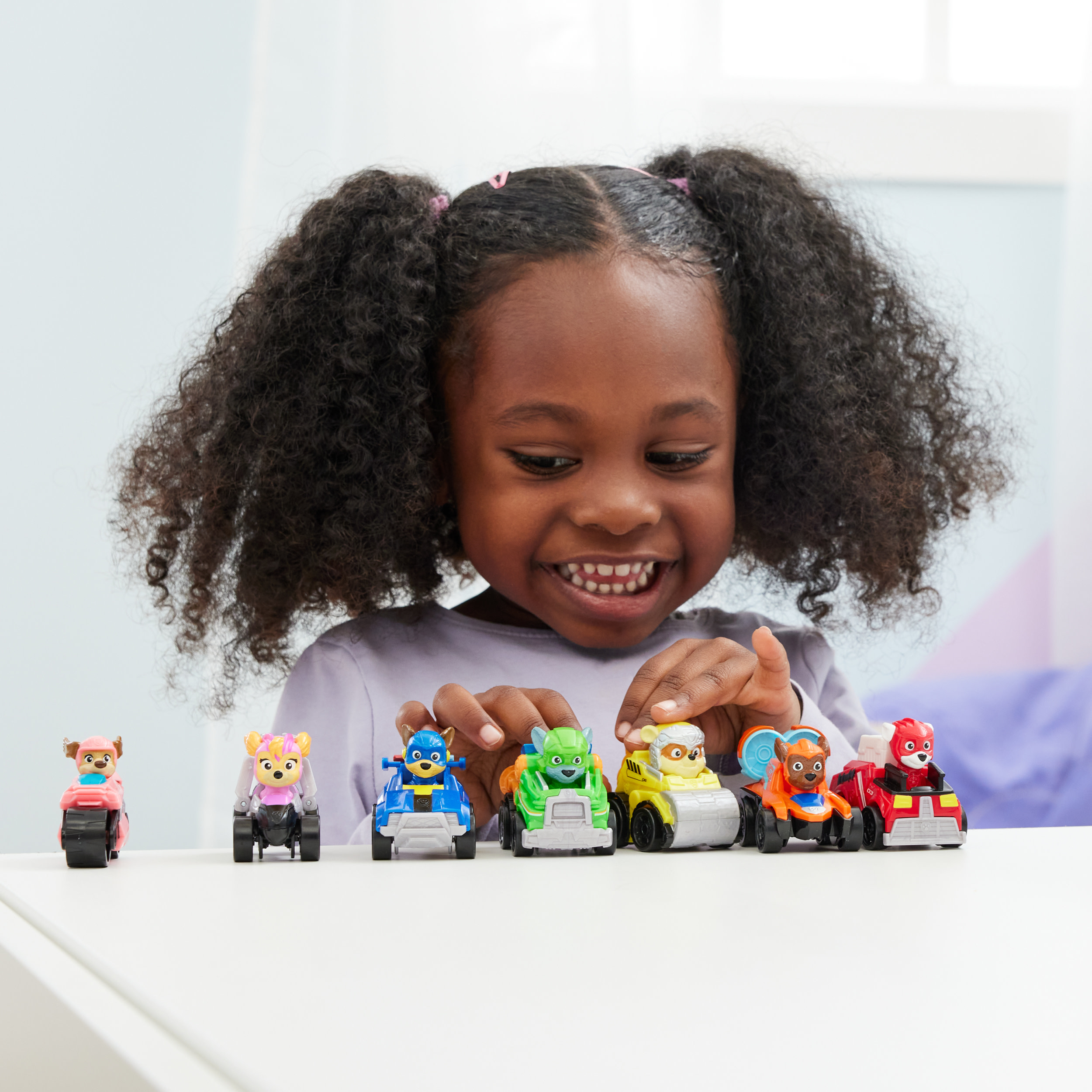 Fille souriant à des voitures jouets Pat' Patrouille. Différentes couleurs : rose, violet, bleu, vert, jaune, orange, rouge.
