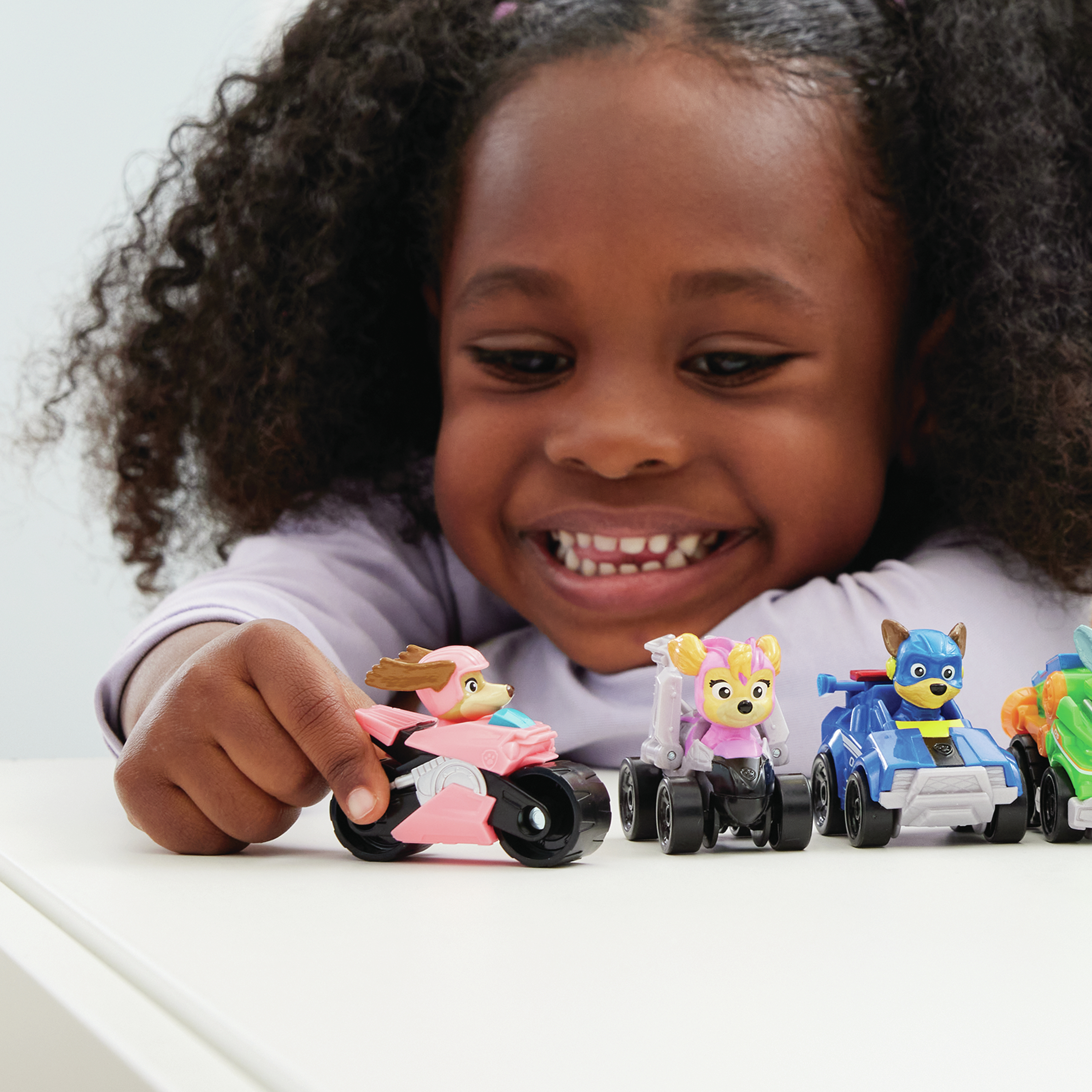 Une fille jouant avec des voitures jouets. Elle sourit et tient une moto rose Pat' Patrouille.