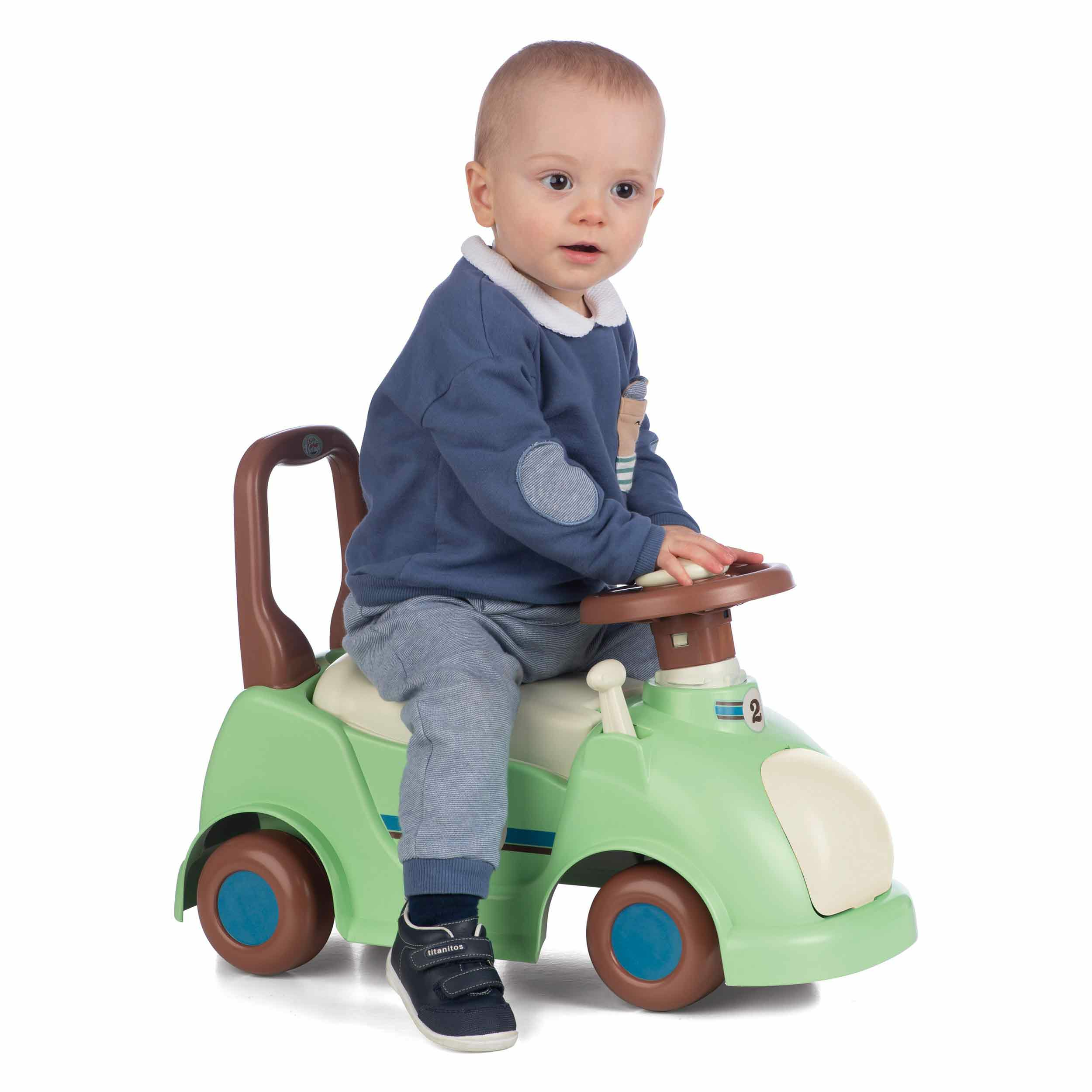 Un bambino è seduto in un'auto giocattolo verde. L'auto ha una maniglia. Il bambino indossa un maglione blu.