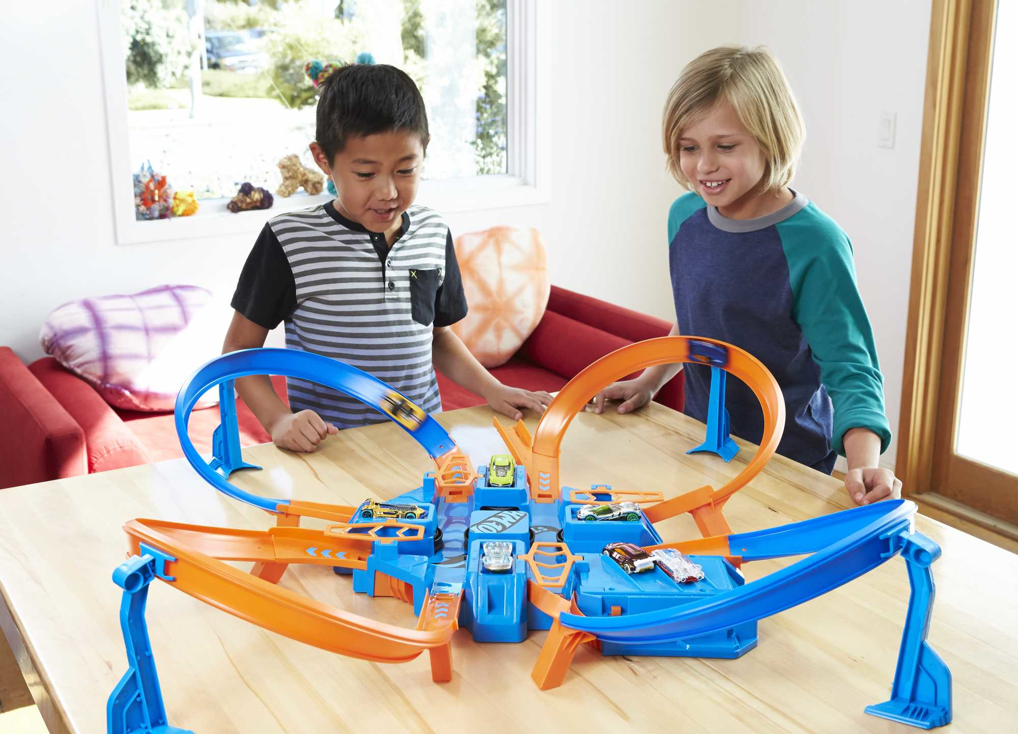 Zwei Kinder spielen mit einem Hot Wheels Rennstrecken-Spielzeugset auf einem Holztisch.