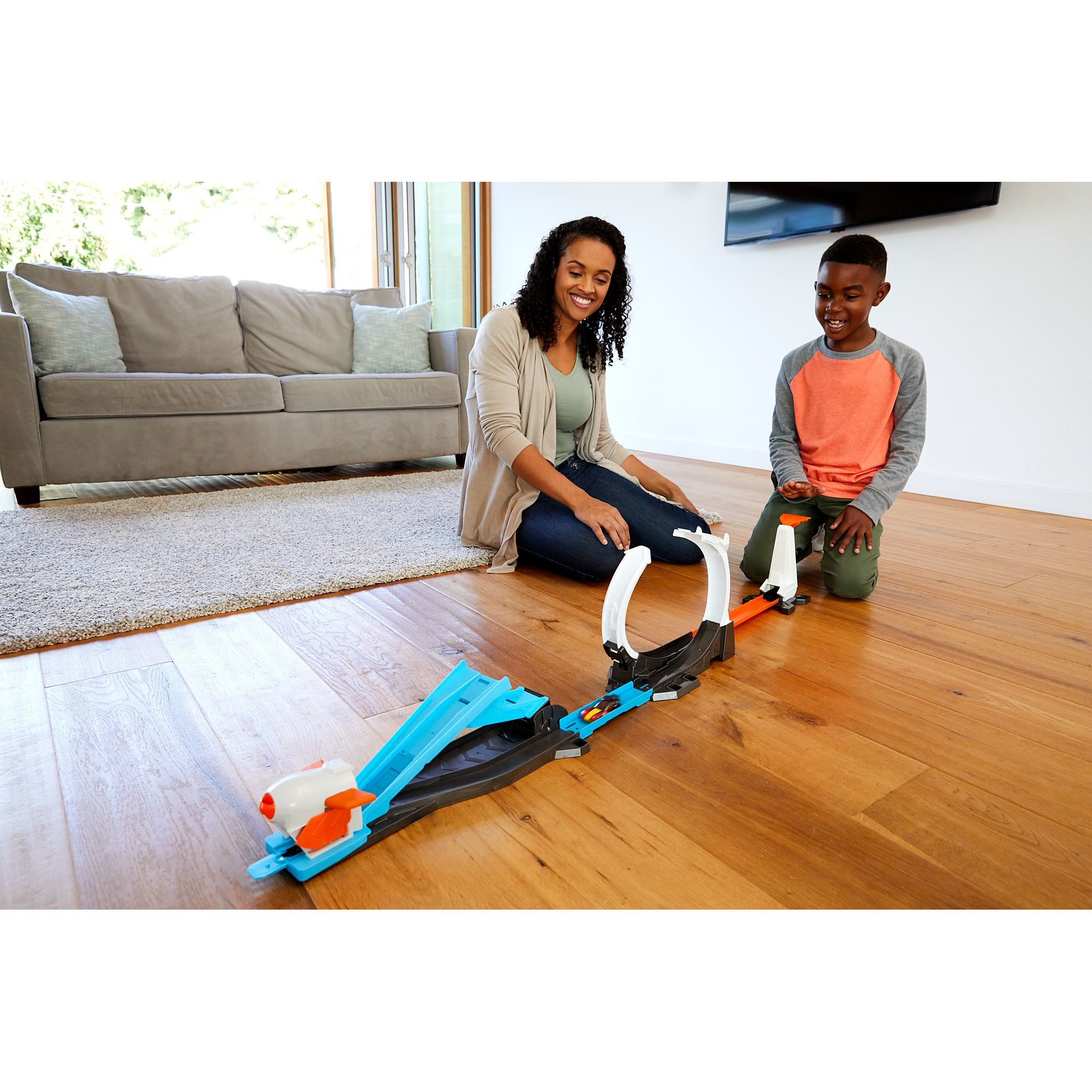 Kobieta i chłopiec bawią się torem samochodowym na drewnianej podłodze. W tle kanapa.