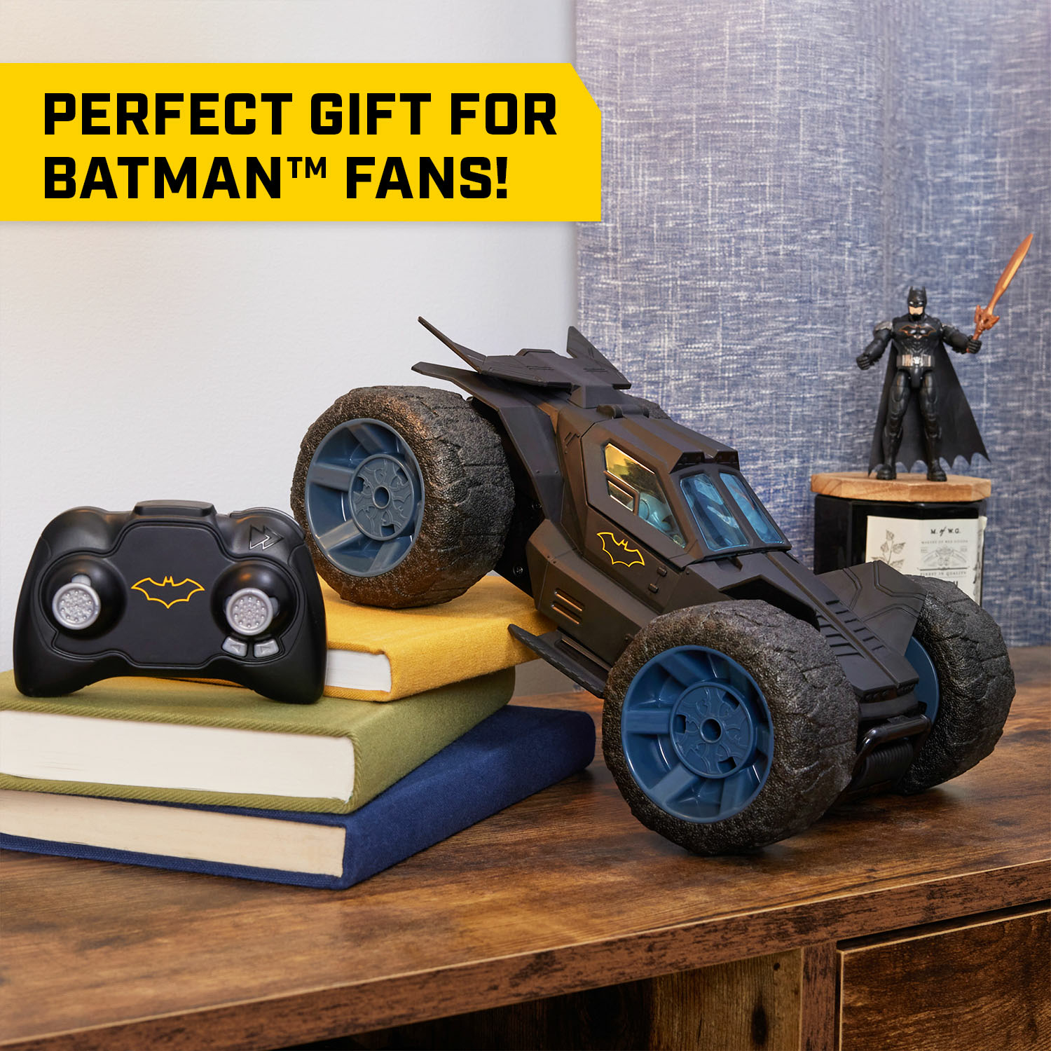 Un coche negro con control remoto y el mando están sobre una mesa con libros. Hay una pequeña figura de Batman cerca.
