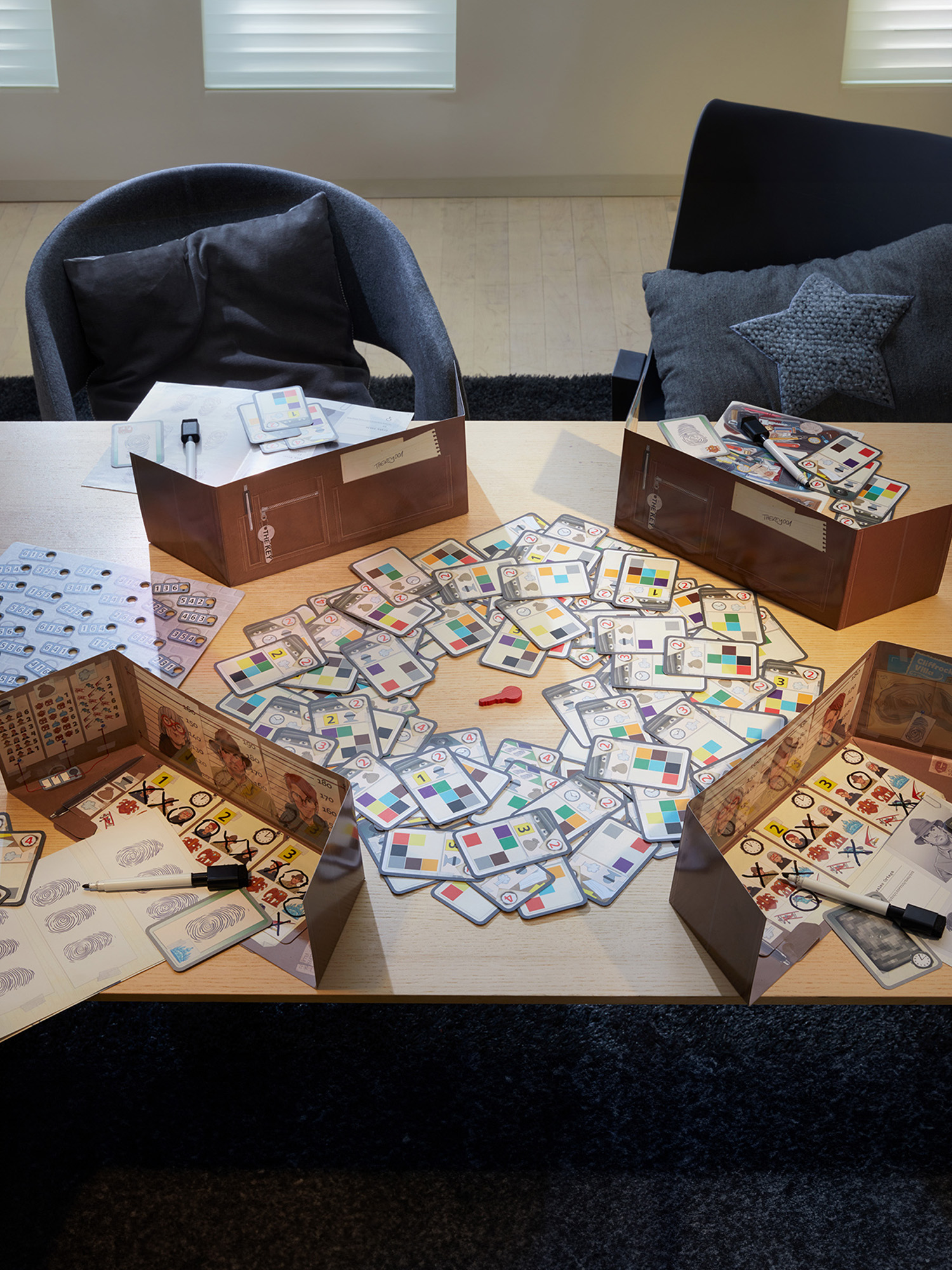 Ein Brettspiel im Spiel auf einem Tisch. Spielsteine, Karten und Schachteln sind angeordnet. Graue Stühle sind sichtbar.