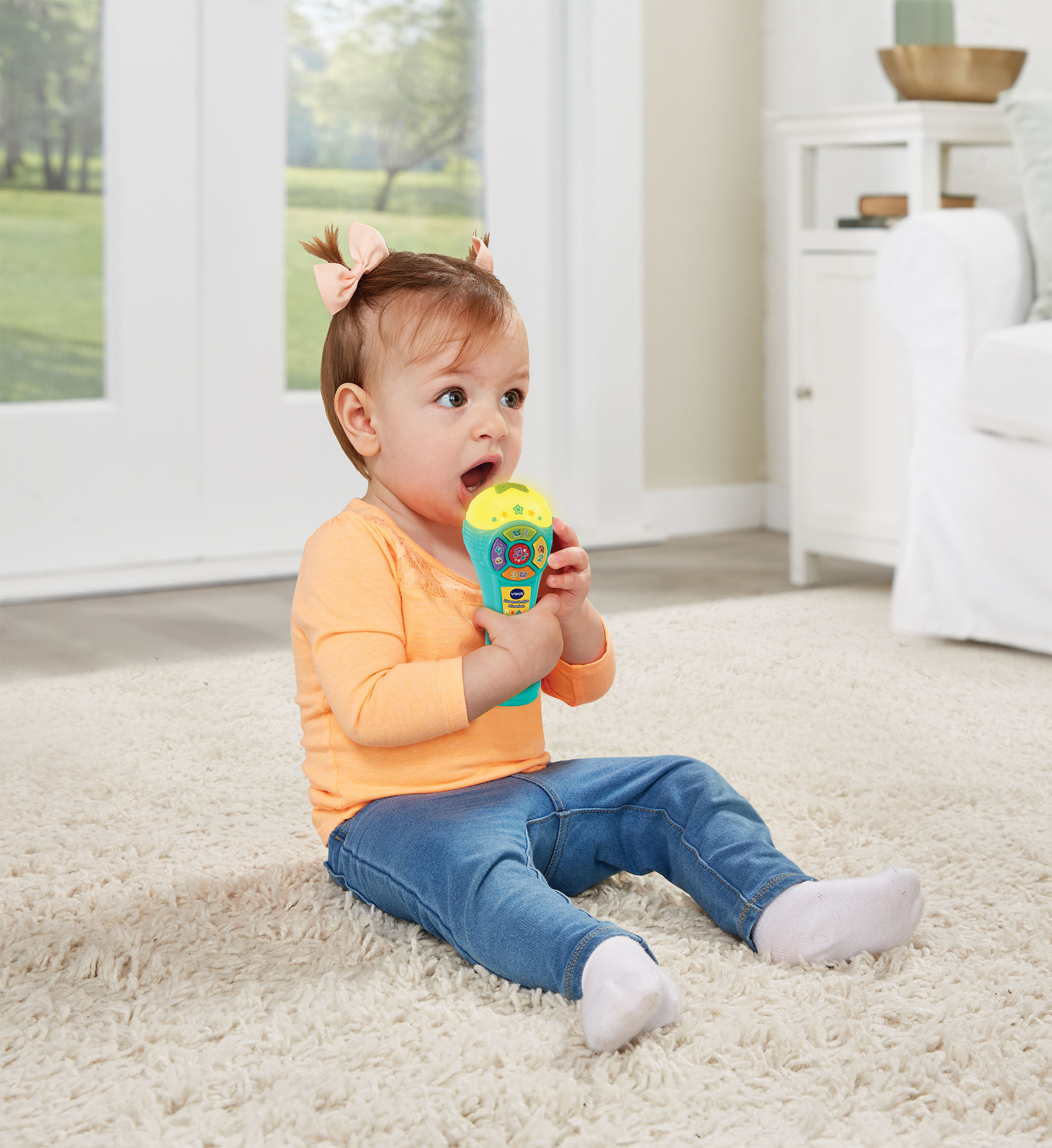 Bambina che tiene in mano un microfono giocattolo turchese. È seduta su un tappeto bianco e indossa una camicia arancione e jeans blu.