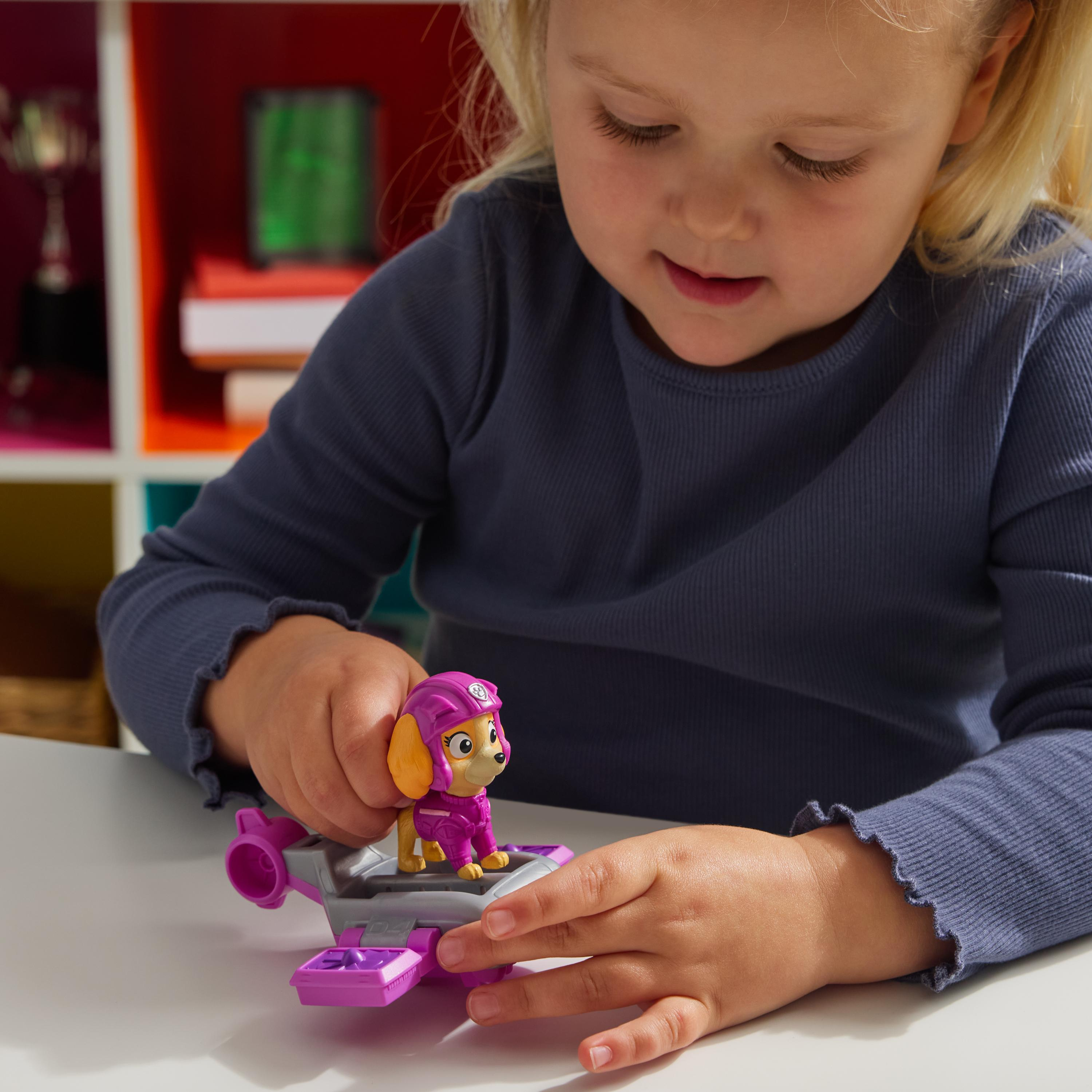 Mädchen spielt mit Skye-Spielzeug und Hoverboard. Weißer Tisch. Bücherregal im Hintergrund. Lächelndes Gesicht.