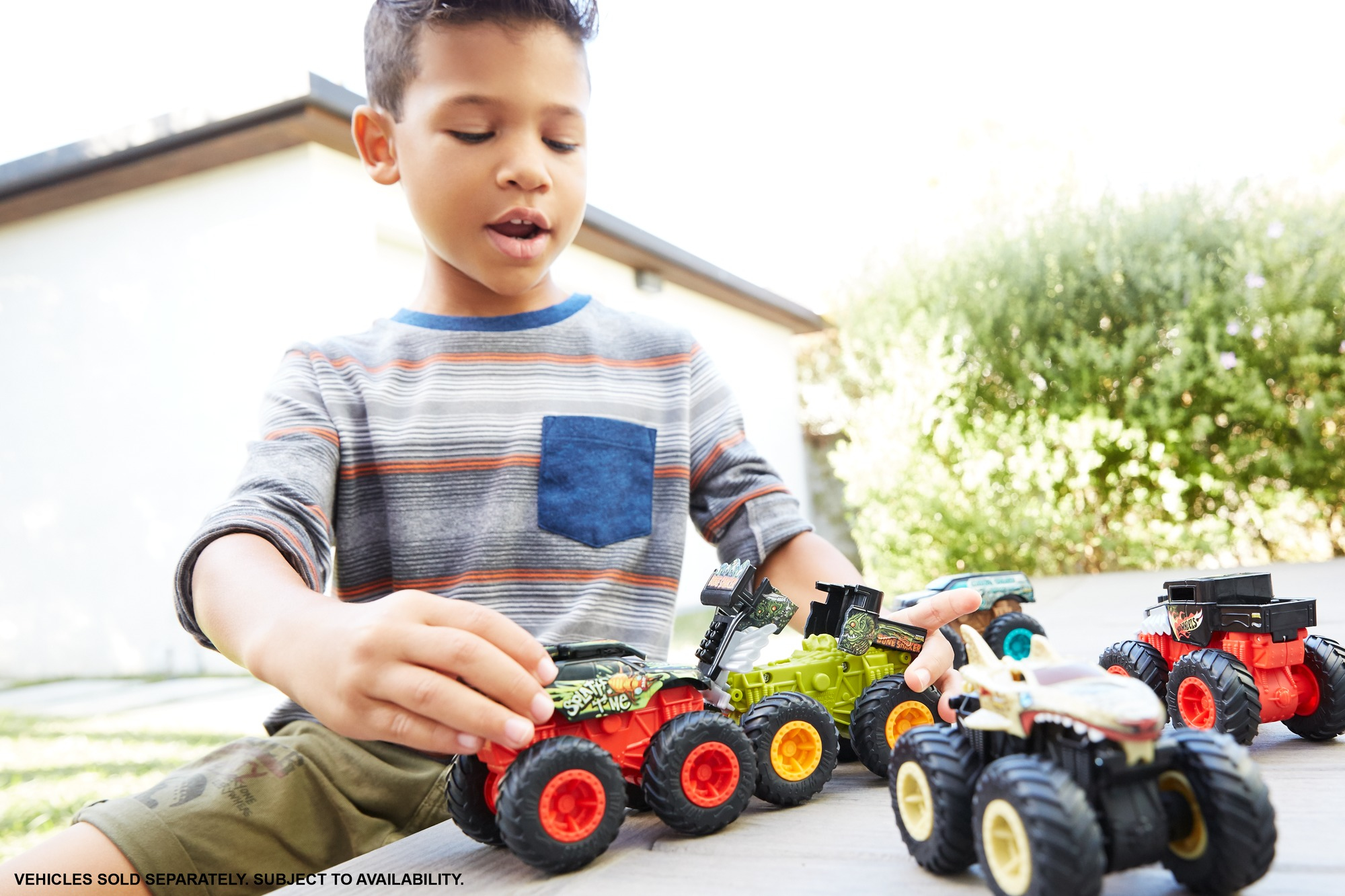 Ein Junge spielt draußen mit Monstertruck-Spielzeugen. Verschiedene bunte Trucks liegen vor ihm auf einem Tisch.