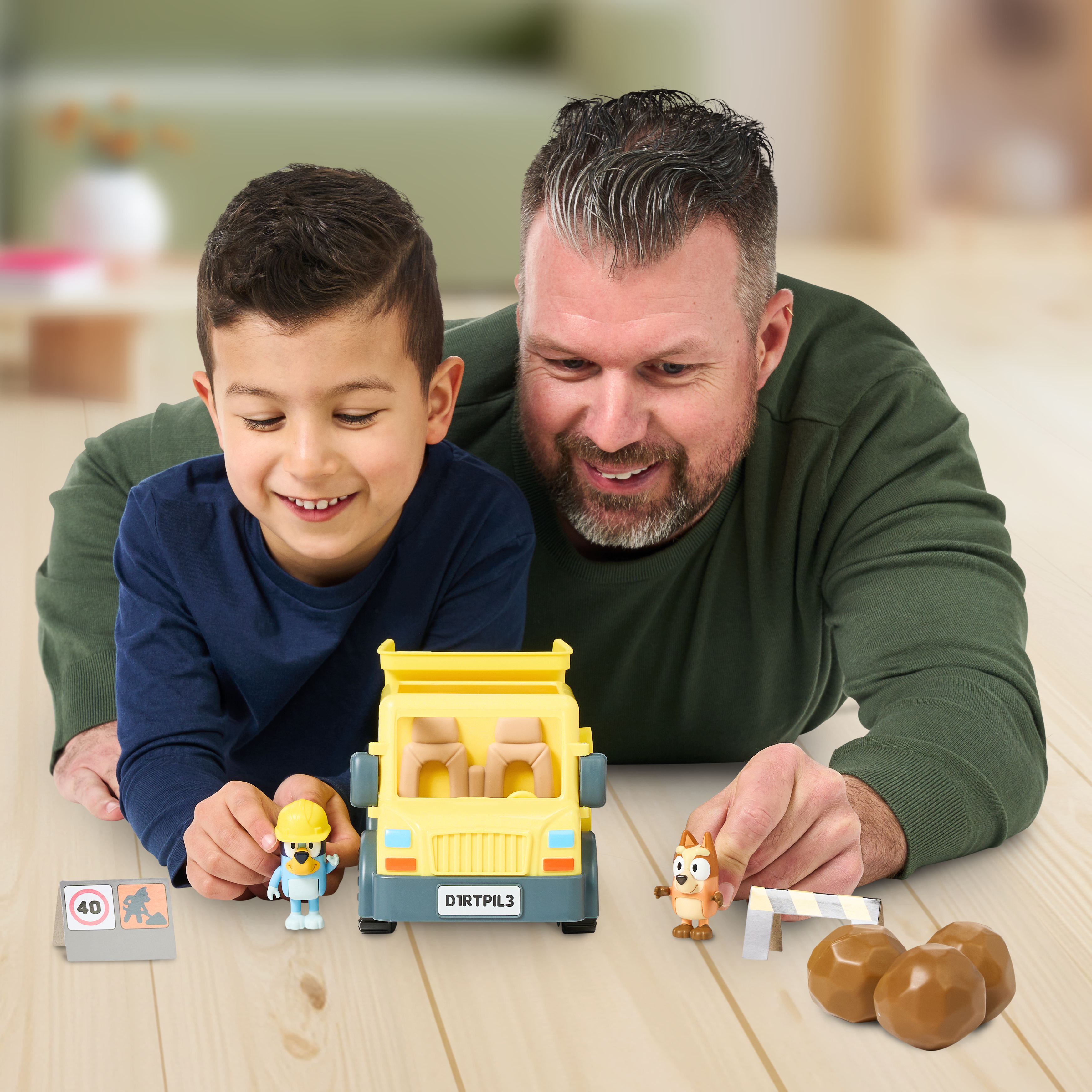 Ojciec i syn bawią się zabawkami, ciężarówkami i figurkami. Scena jest w pomieszczeniu.