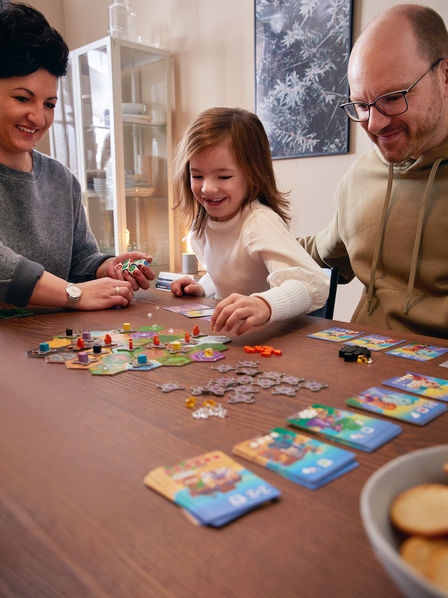 Eine Familie spielt ein Brettspiel an einem braunen Tisch, lächelt, mit Karten und Spielsteinen.