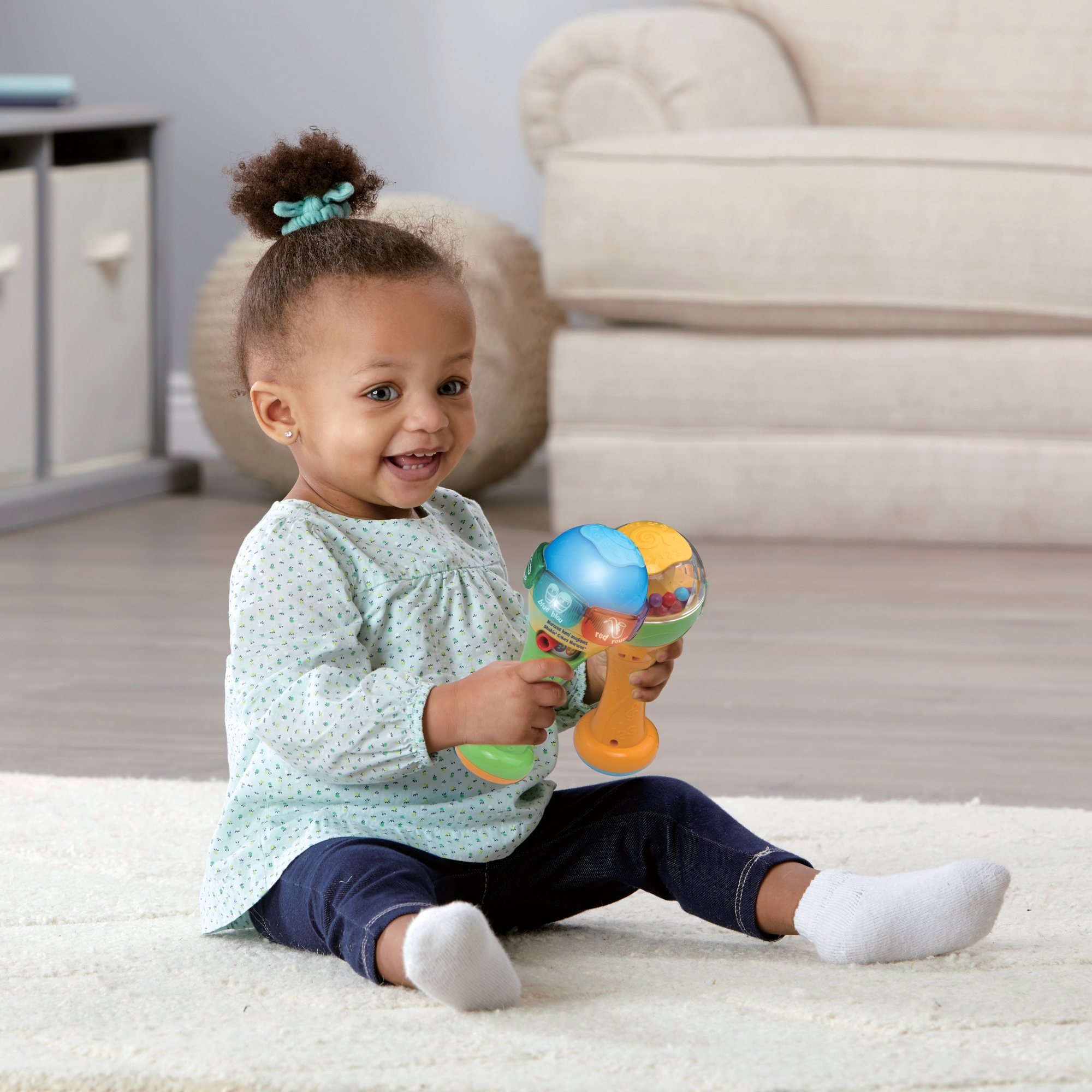 Lachendes Baby hält eine bunte Rassel. Das Baby sitzt auf einem weißen Teppich und sieht glücklich aus.