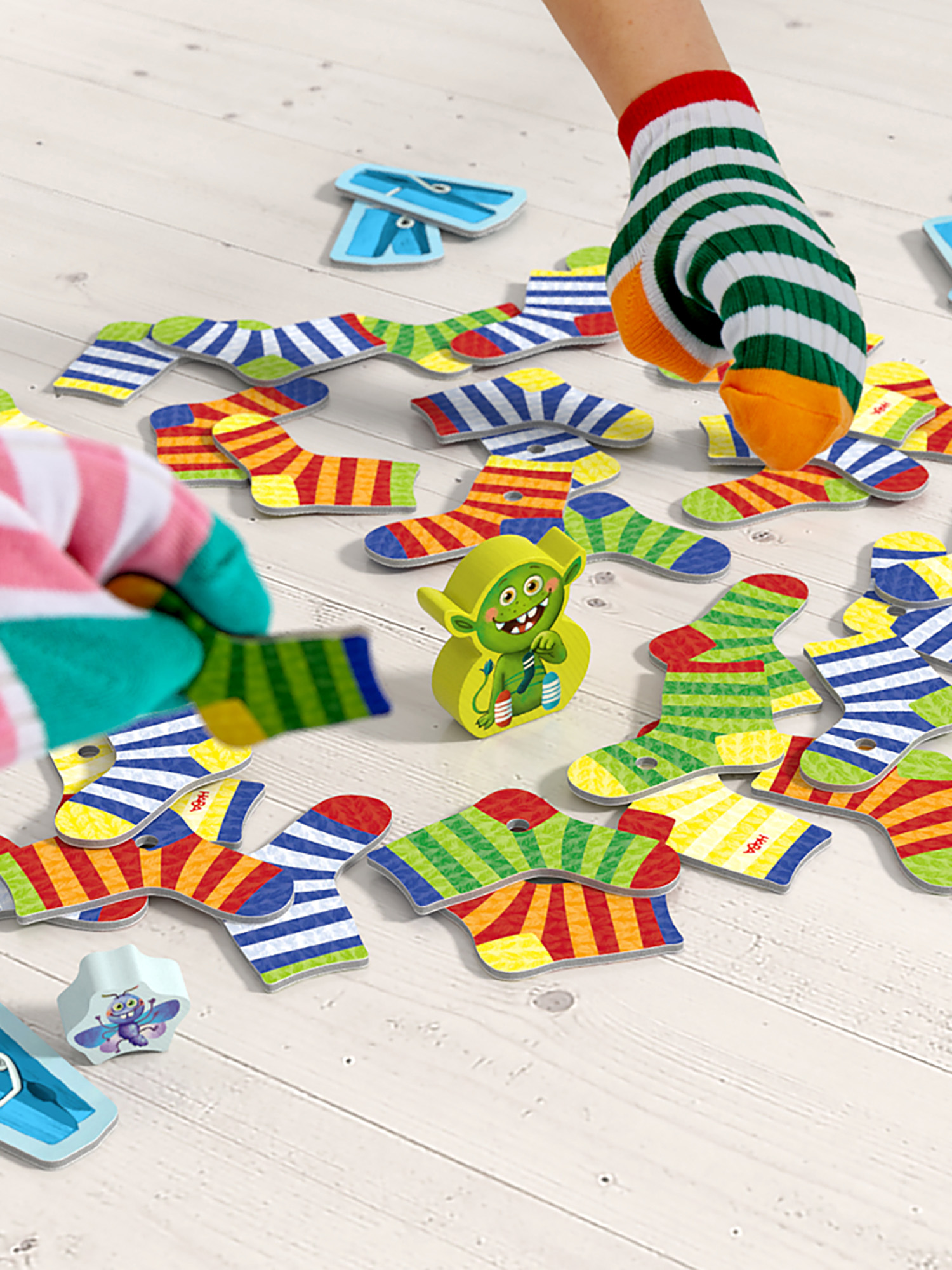 Hände mit gestreiften Socken spielen ein Brettspiel mit bunten sockenförmigen Spielsteinen und einem grünen Monster.