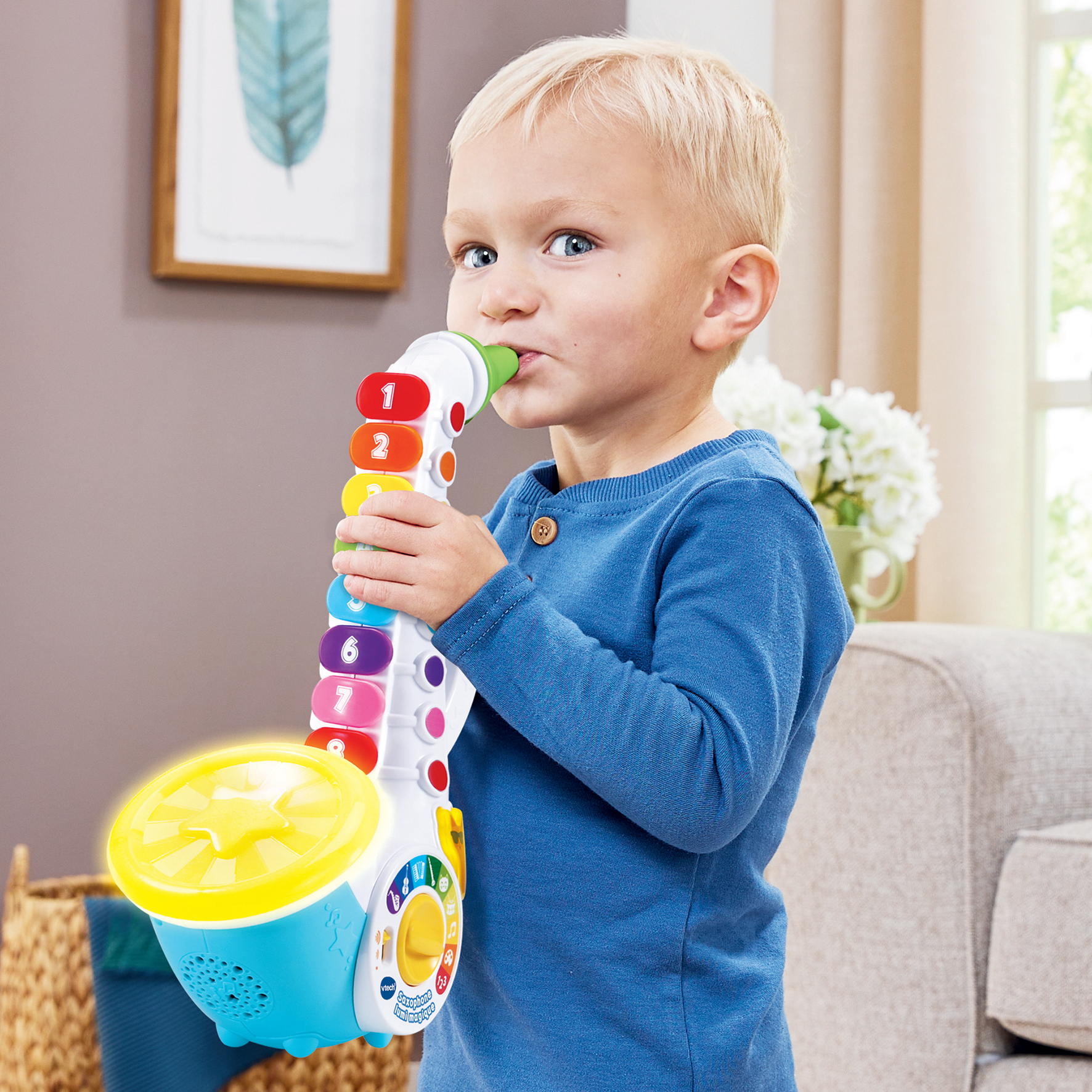 Niño rubio tocando un saxofón de juguete colorido en interiores, con un cuadro en el fondo.