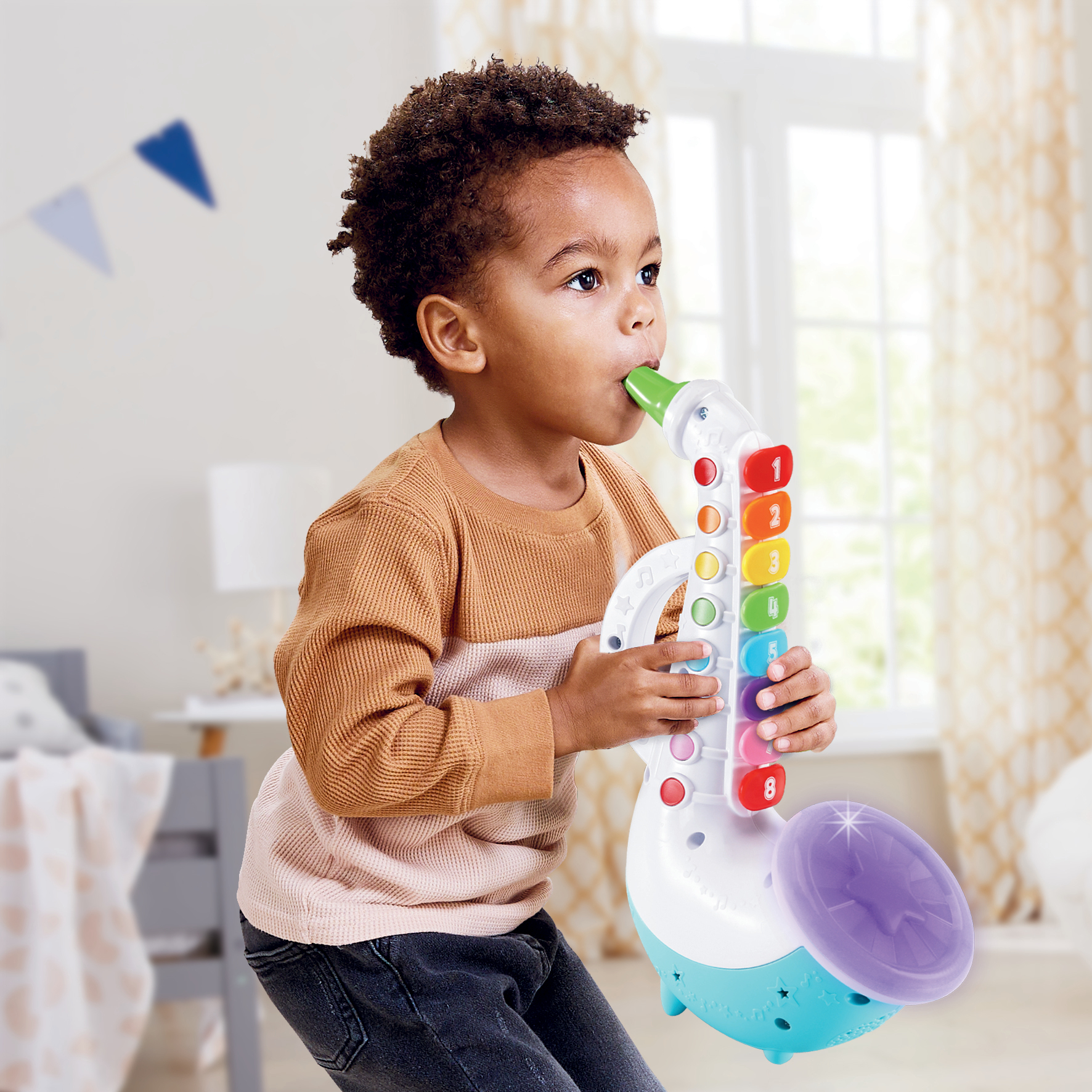 Niño pequeño tocando un saxofón de juguete colorido en interiores, con una ventana y una habitación neutra.