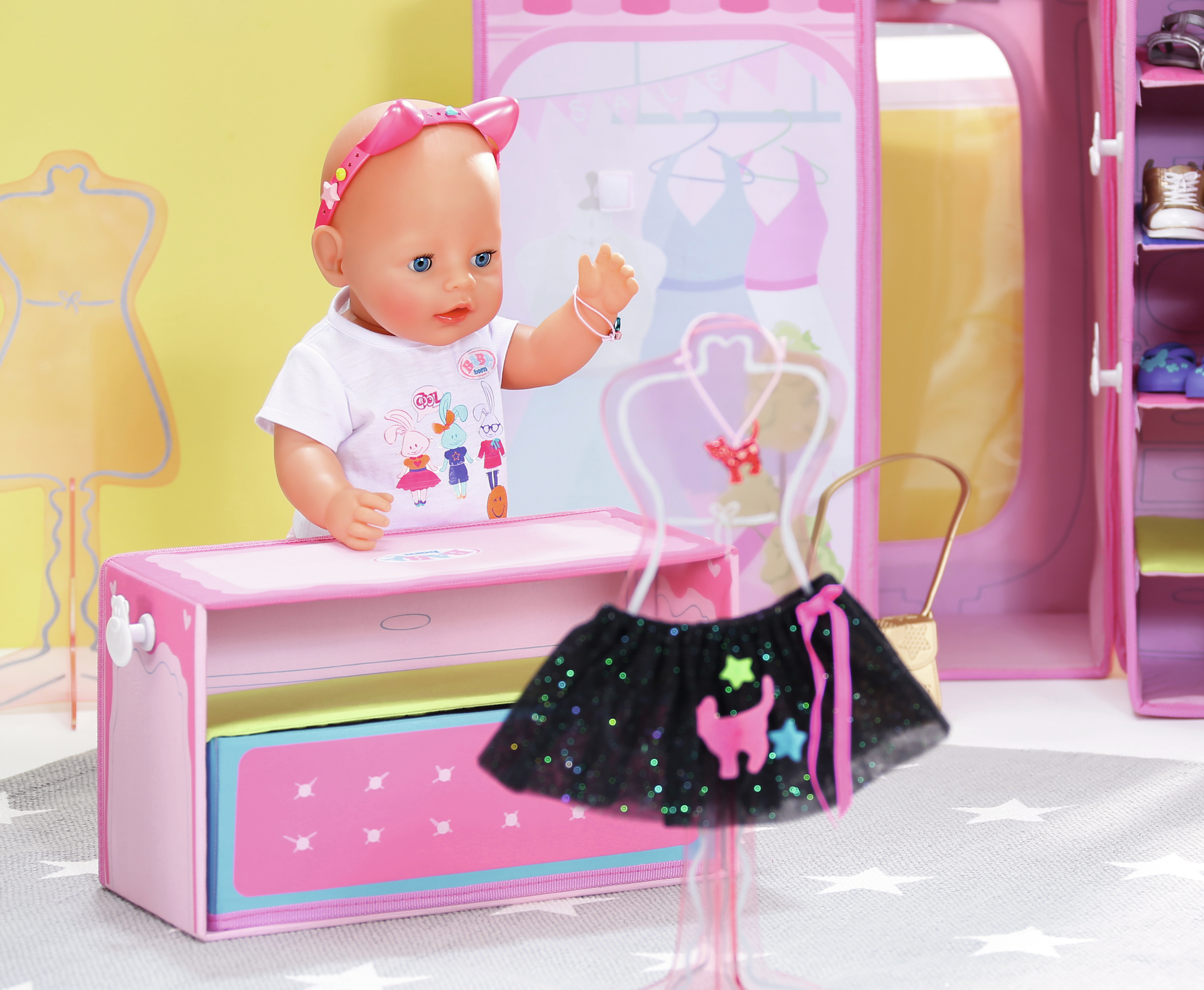 Lalka stoi obok szafy. Lalka ma różową opaskę na głowę i biały t-shirt. Spódnica jest wystawiona.