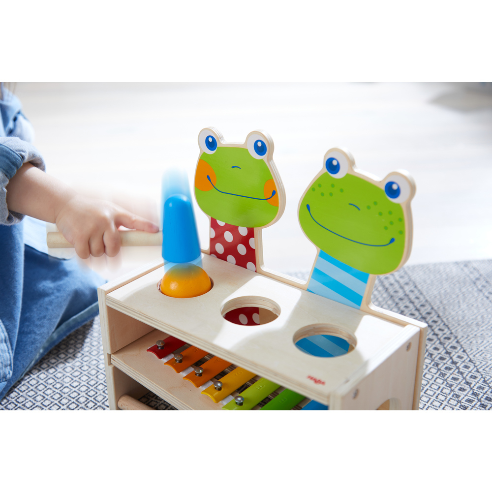 Holzspielzeug mit zwei Froschgesichtern. Eine Hand schlägt mit dem Hammer. Weißer Hintergrund.