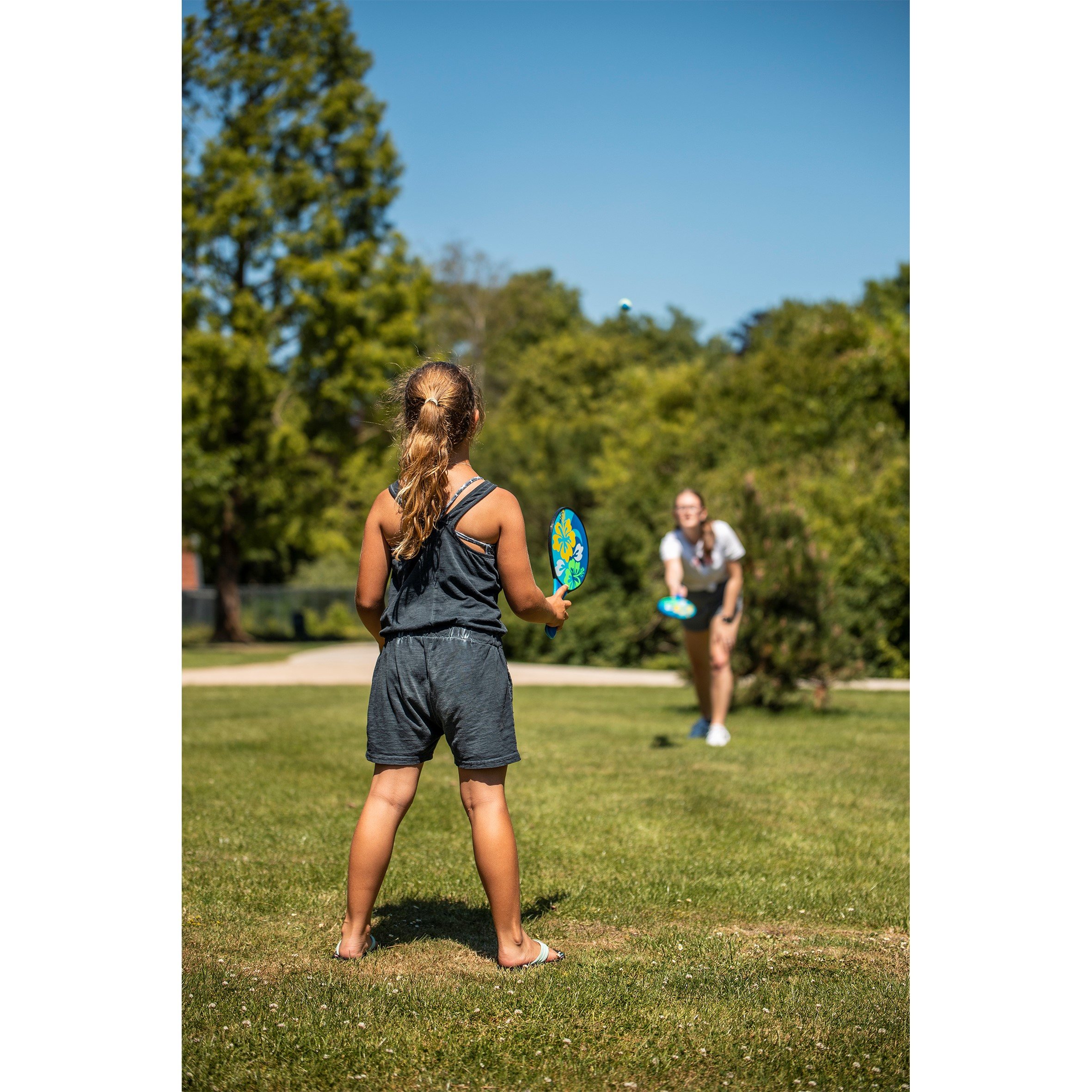 Ragazza che gioca a paddleball sulla spiaggia. Un'altra ragazza sullo sfondo. Erba verde.
