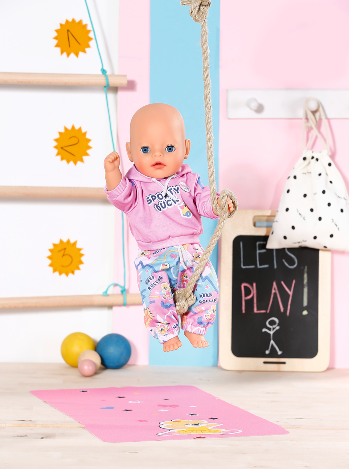 Lalka wisi na linie, ubrana w różową bluzę z kapturem i spodnie, pod spodem różowa mata do jogi.