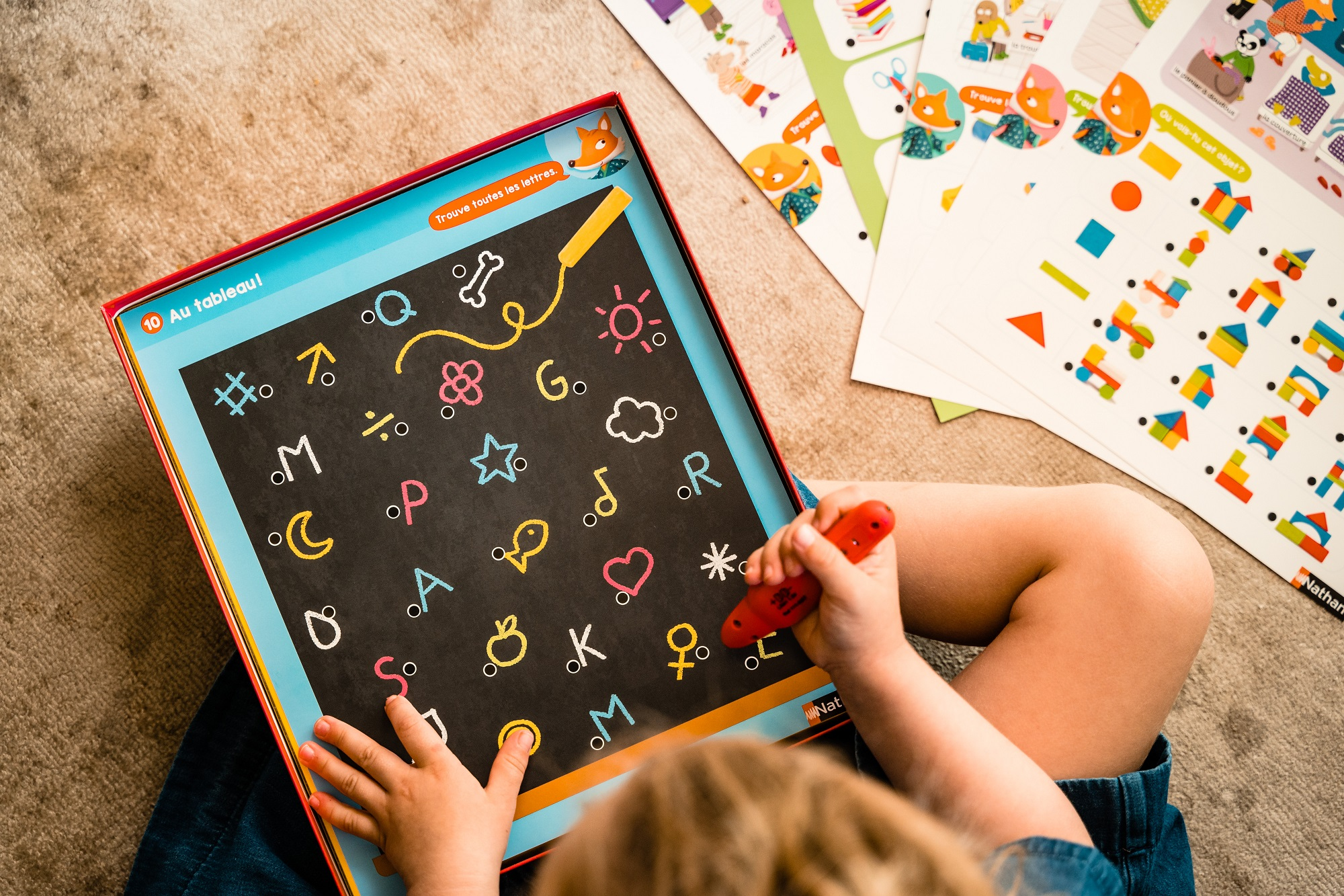 Dziecko bawi się grą planszową, wskazując czerwonym długopisem litery, kształty i symbole.