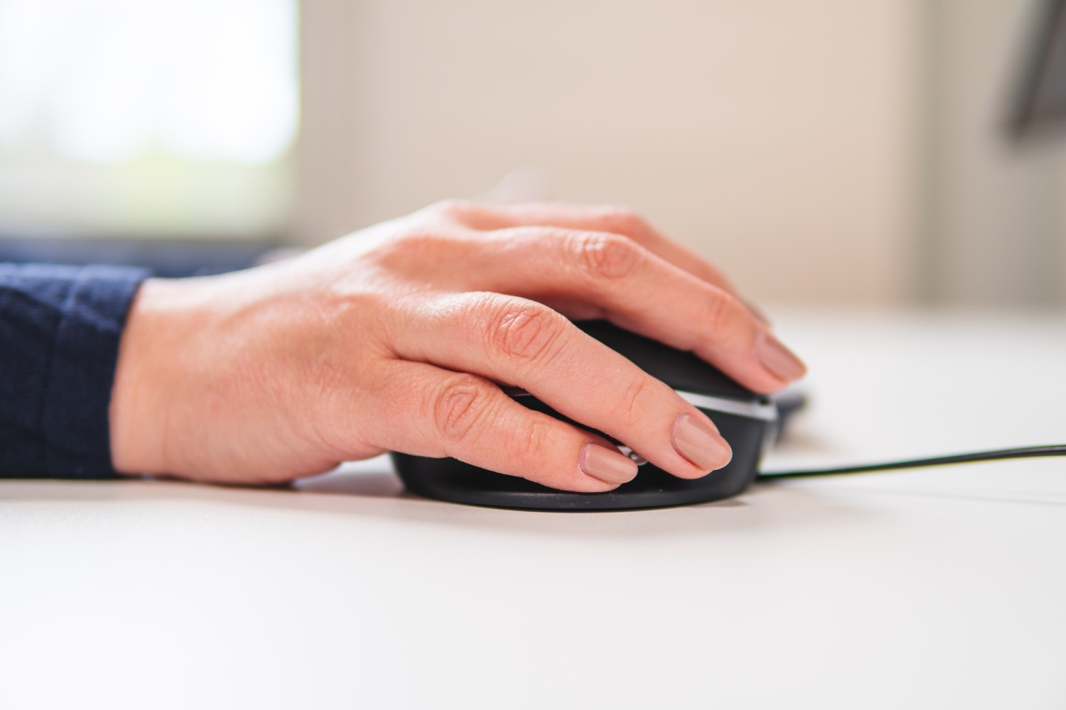 Une main posée sur une souris d'ordinateur noire sur un bureau blanc. Les ongles sont vernis.