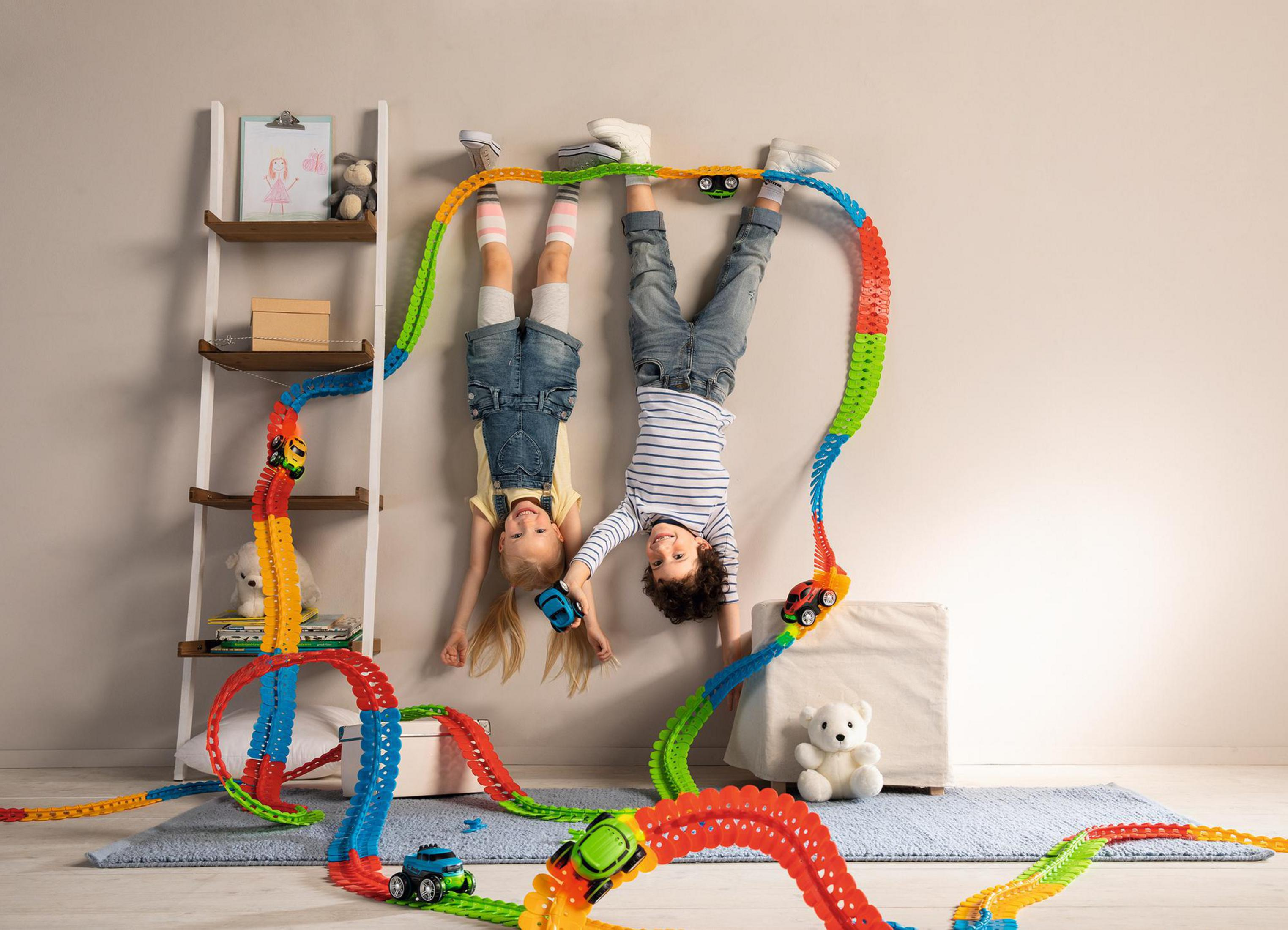 Dwoje dzieci zwisa do góry nogami z toru zabawek. Zabawki samochodowe są na torze.