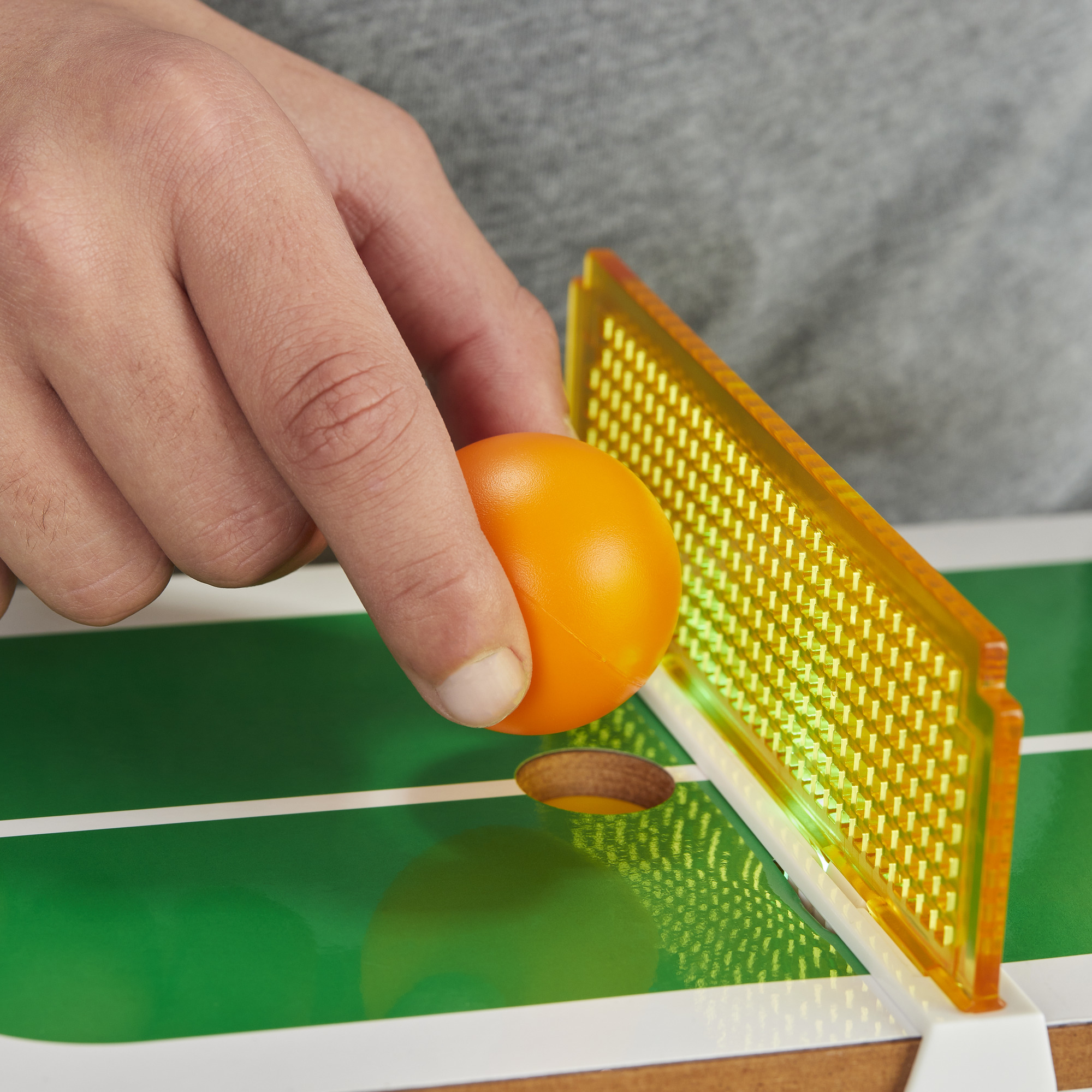 Una mano che tiene una pallina da ping pong arancione vicino a una rete e una superficie verde.