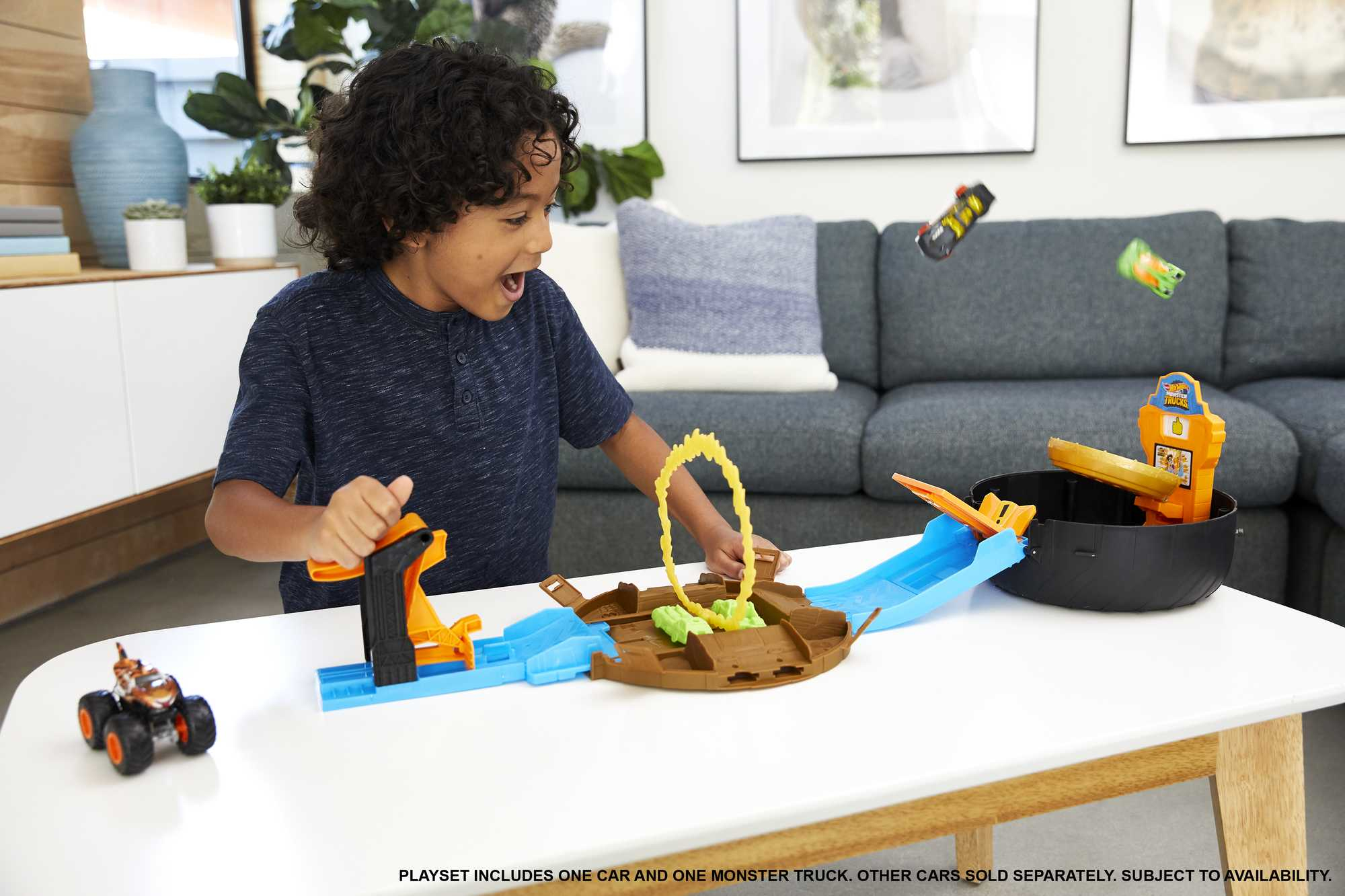 Chłopiec bawi się torem Hot Wheels. Samochody latają. Biały stół z szarą sofą w tle.