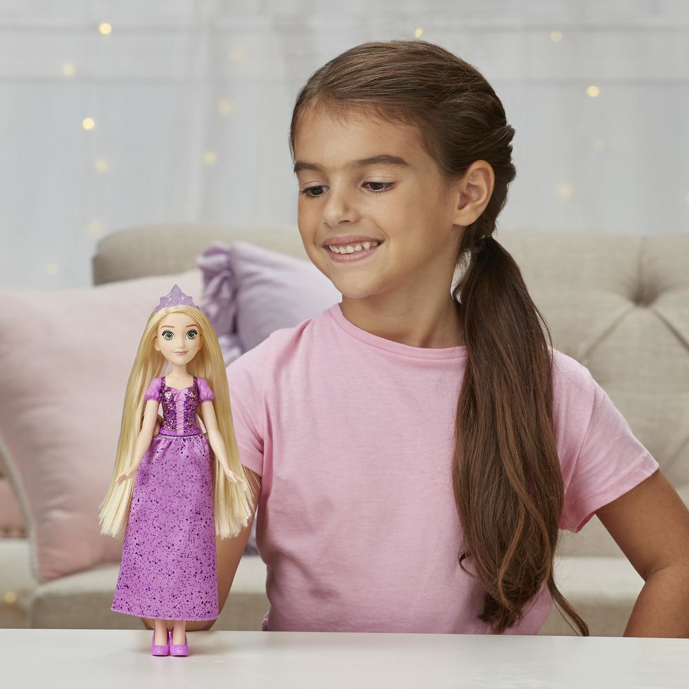 Ragazza sorridente, tiene in mano la bambola di Rapunzel. Capelli biondi, vestito viola. Sfondo: divano e cuscini.