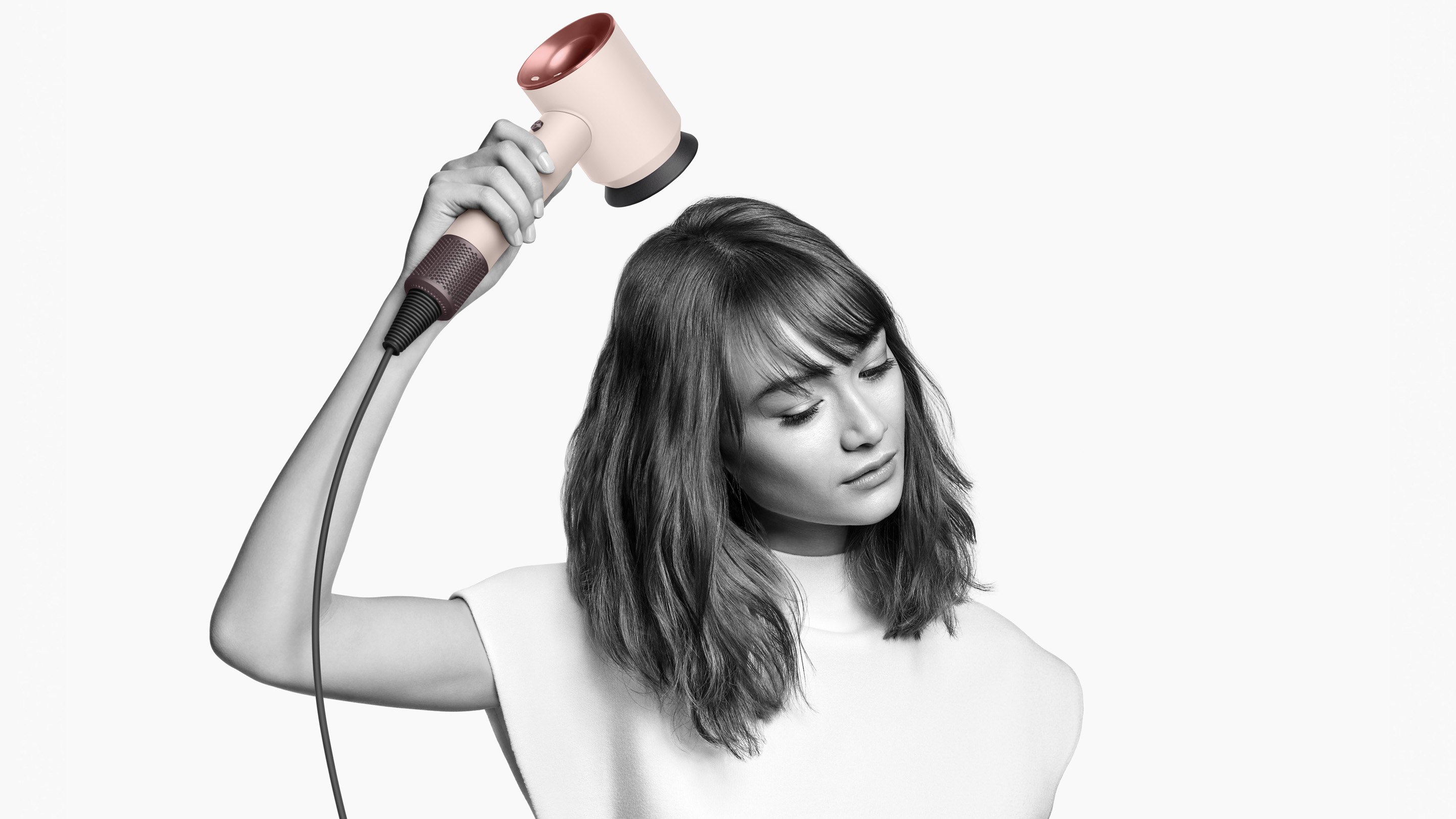 Beyaz ve gül altın rengi bir Dyson saç kurutma makinesi kullanan bir kadın. Kurutma makinesini saçının üzerinde tutuyor.