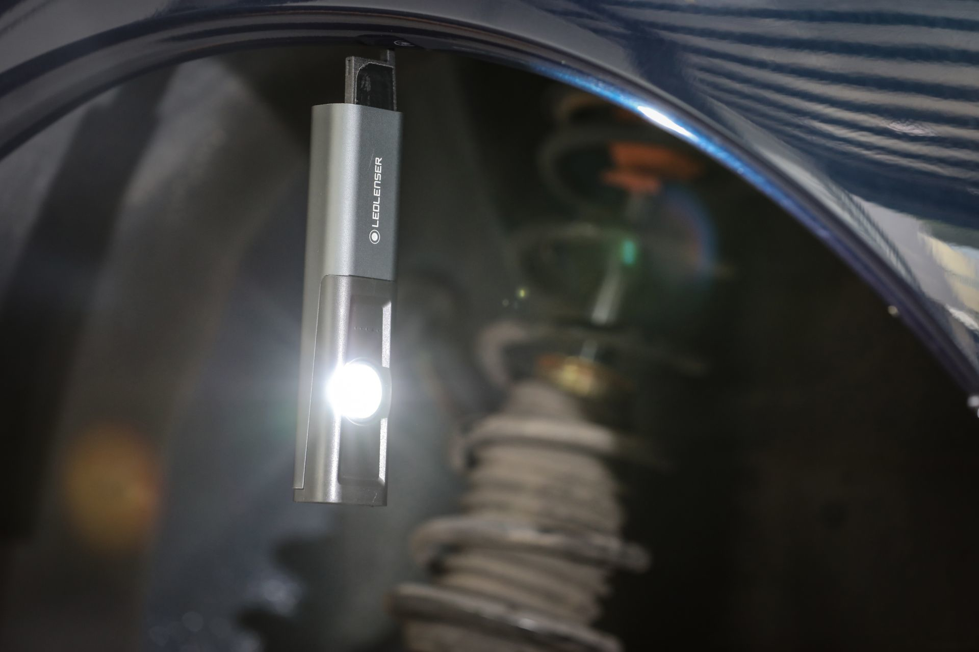 Srebrna latarka LEDLENSER świeci światłem pod niebieskim błotnikiem samochodowym.