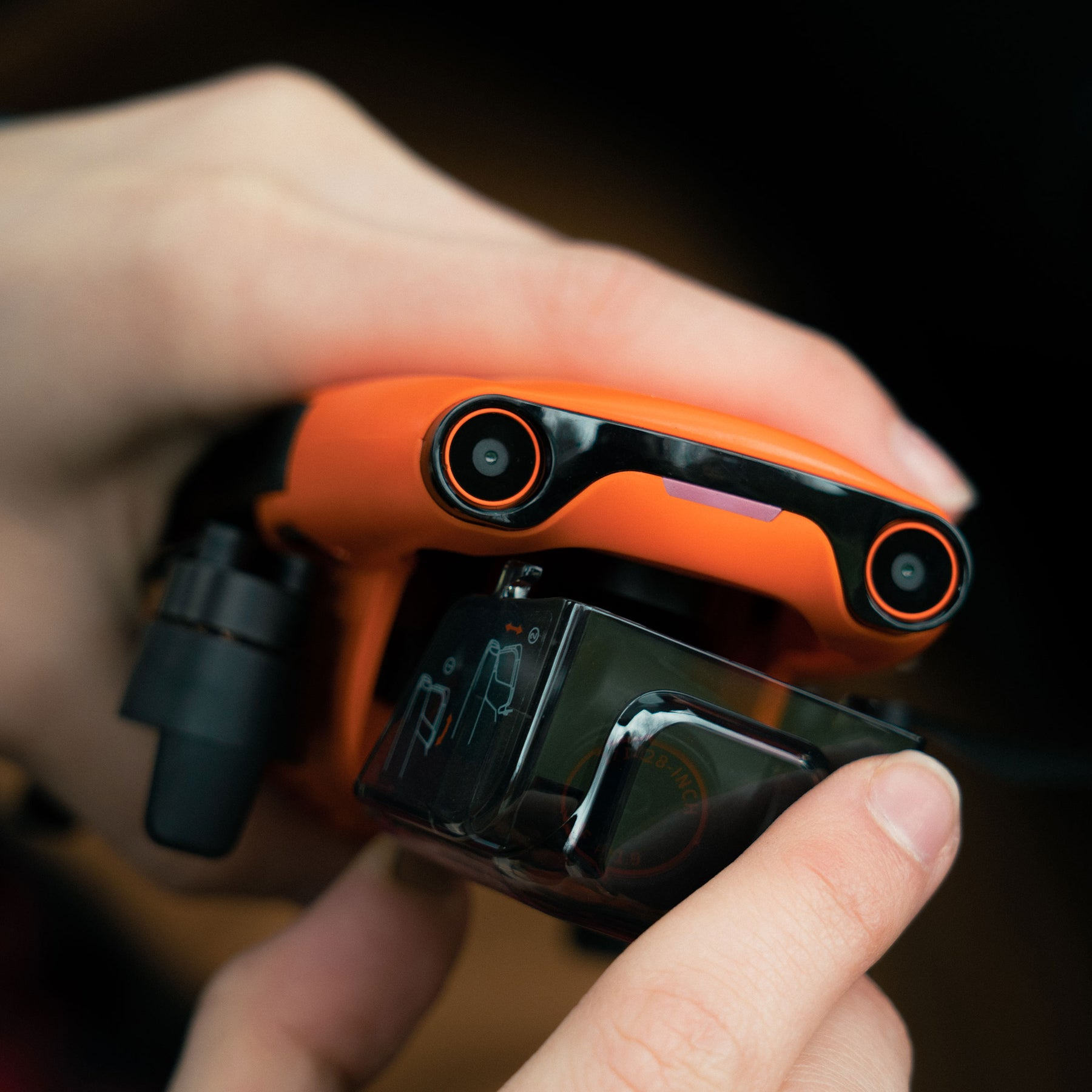 Mani che tengono un drone arancione. Il drone ha due fotocamere e una batteria nera.
