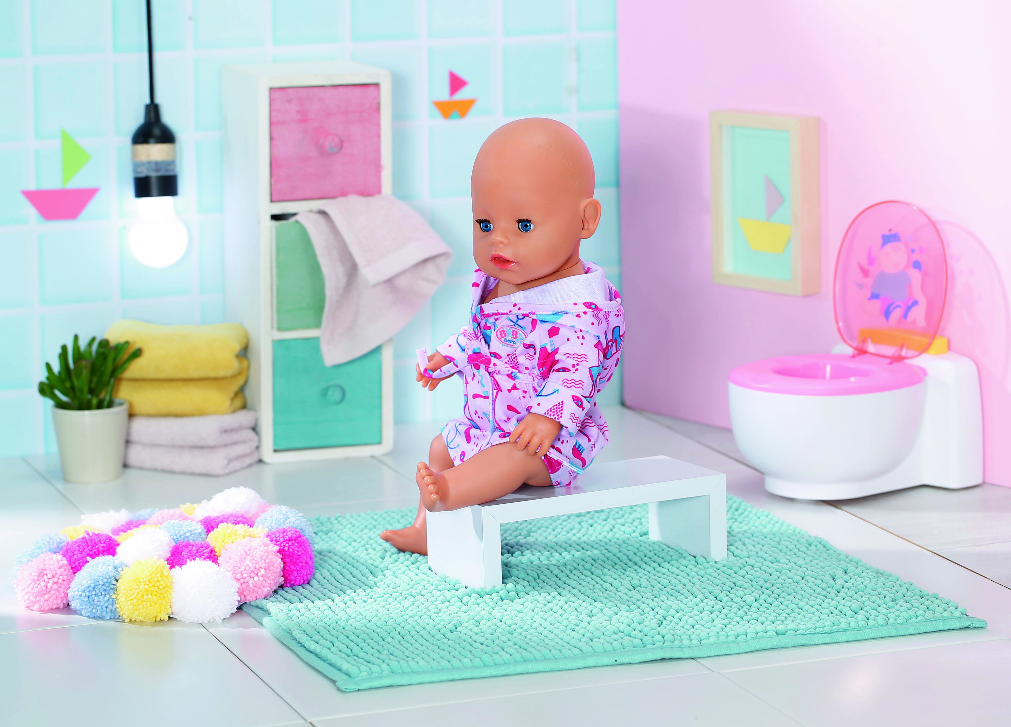 Lalka w łazience siedząca na białej ławce, w szlafroku. Jest toaleta i mata.