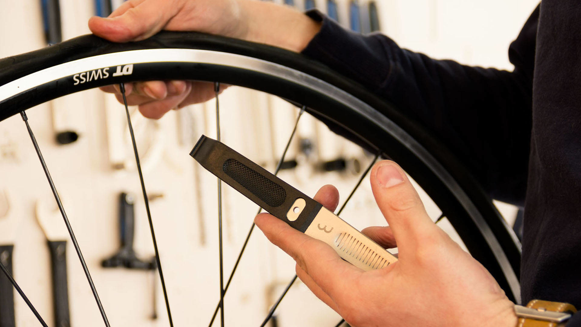 Dłonie trzymające łyżkę do opon, obok opony rowerowej i koła, w warsztacie.