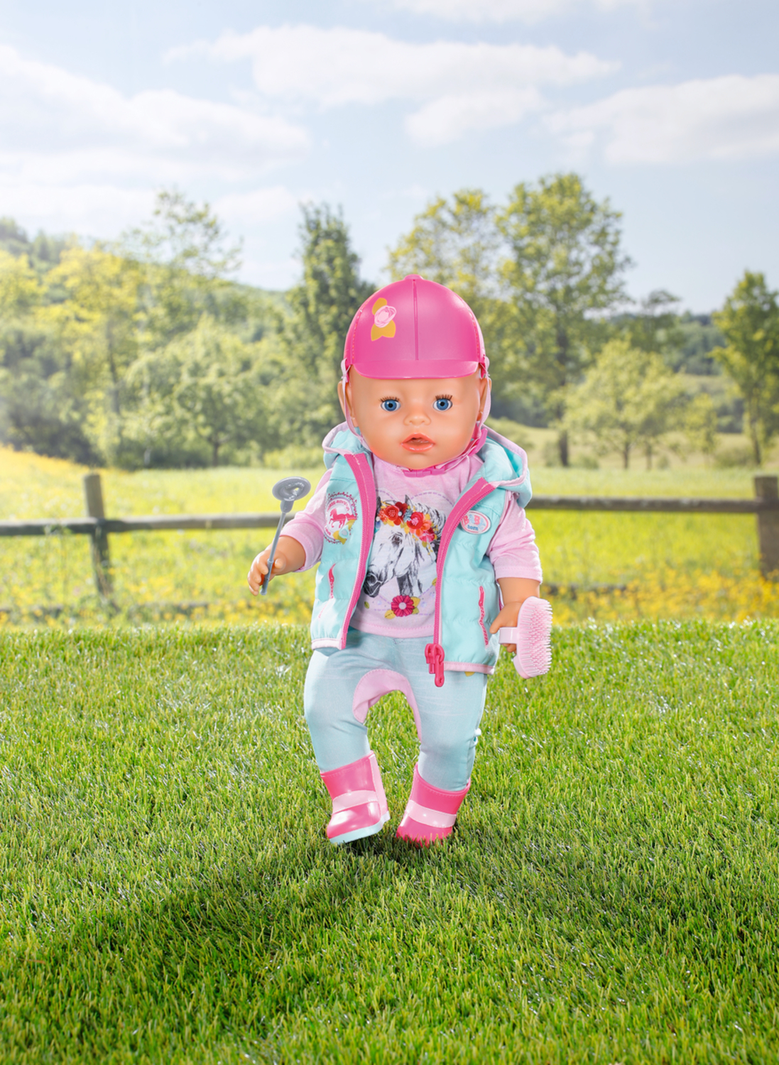 Lalka w stroju jeździeckim stoi na trawie, trzymając palcat i szczotkę, w różowym kasku.