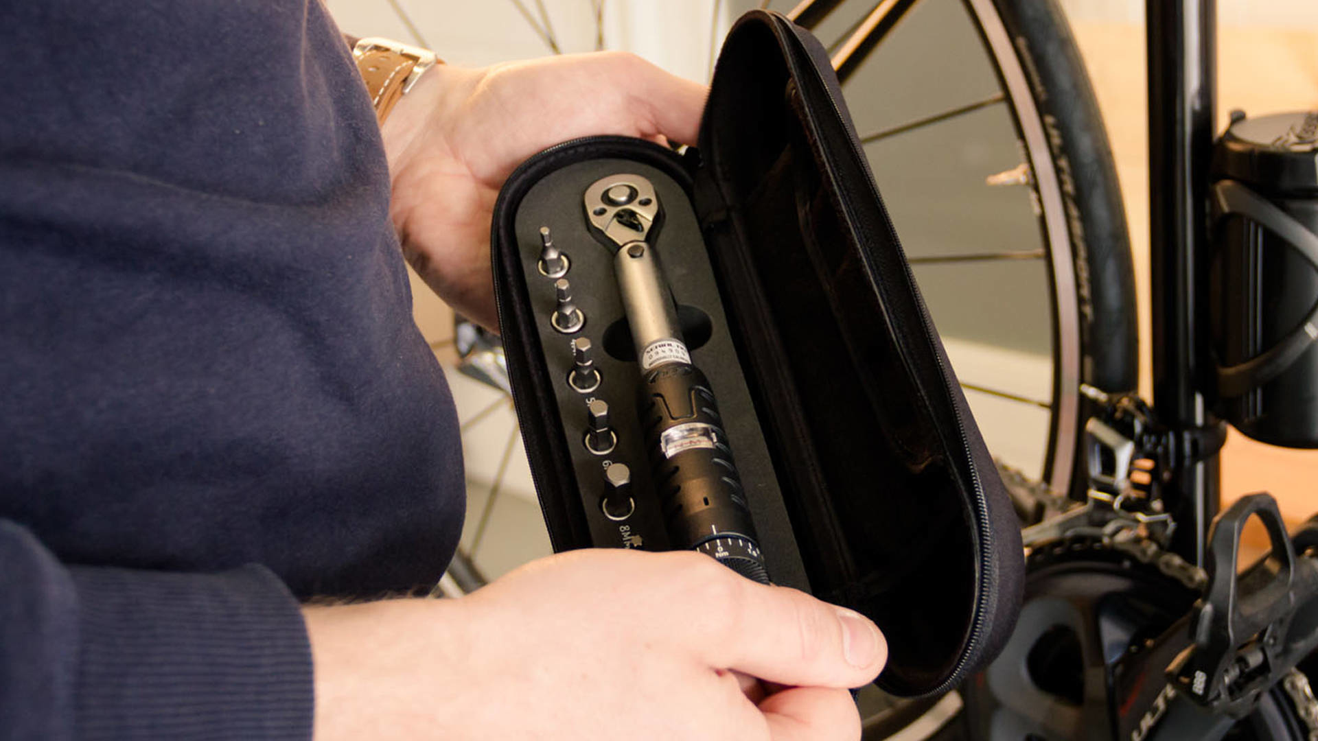 Osoba trzymająca otwartą czarną walizkę z kluczem dynamometrycznym i bitami, rower w tle.