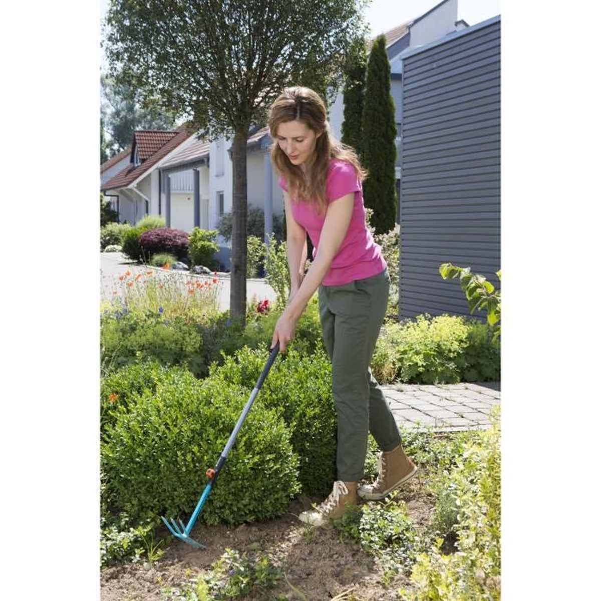 Frau harkt Erde in einem Garten mit grünen Büschen, Häuser im Hintergrund.