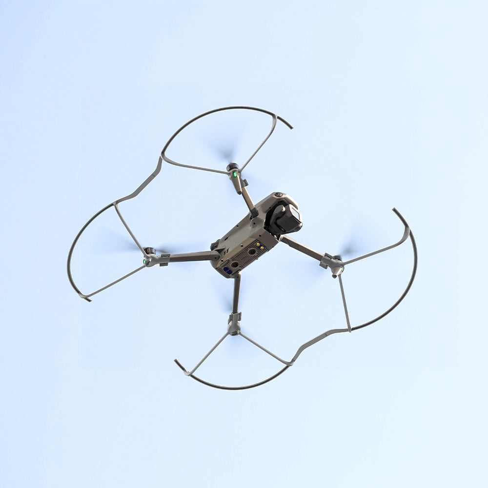 Dron na niebie, cztery śmigła, szary korpus, niebieskie tło.