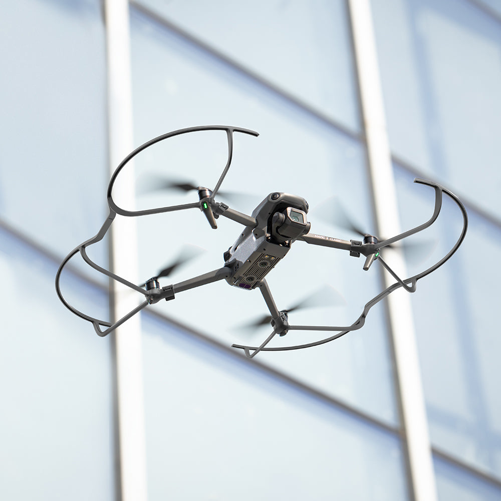 Dron w locie, z osłonami śmigieł, na tle szklanego budynku. Dron jest szary i czarny.