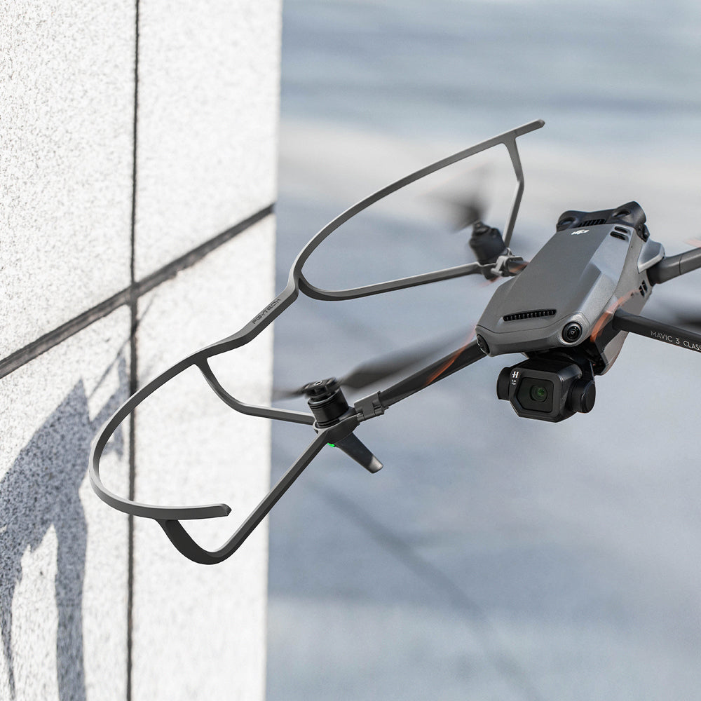 Dron w locie z ramą ochronną. Szare i pomarańczowe detale na tle budynku.