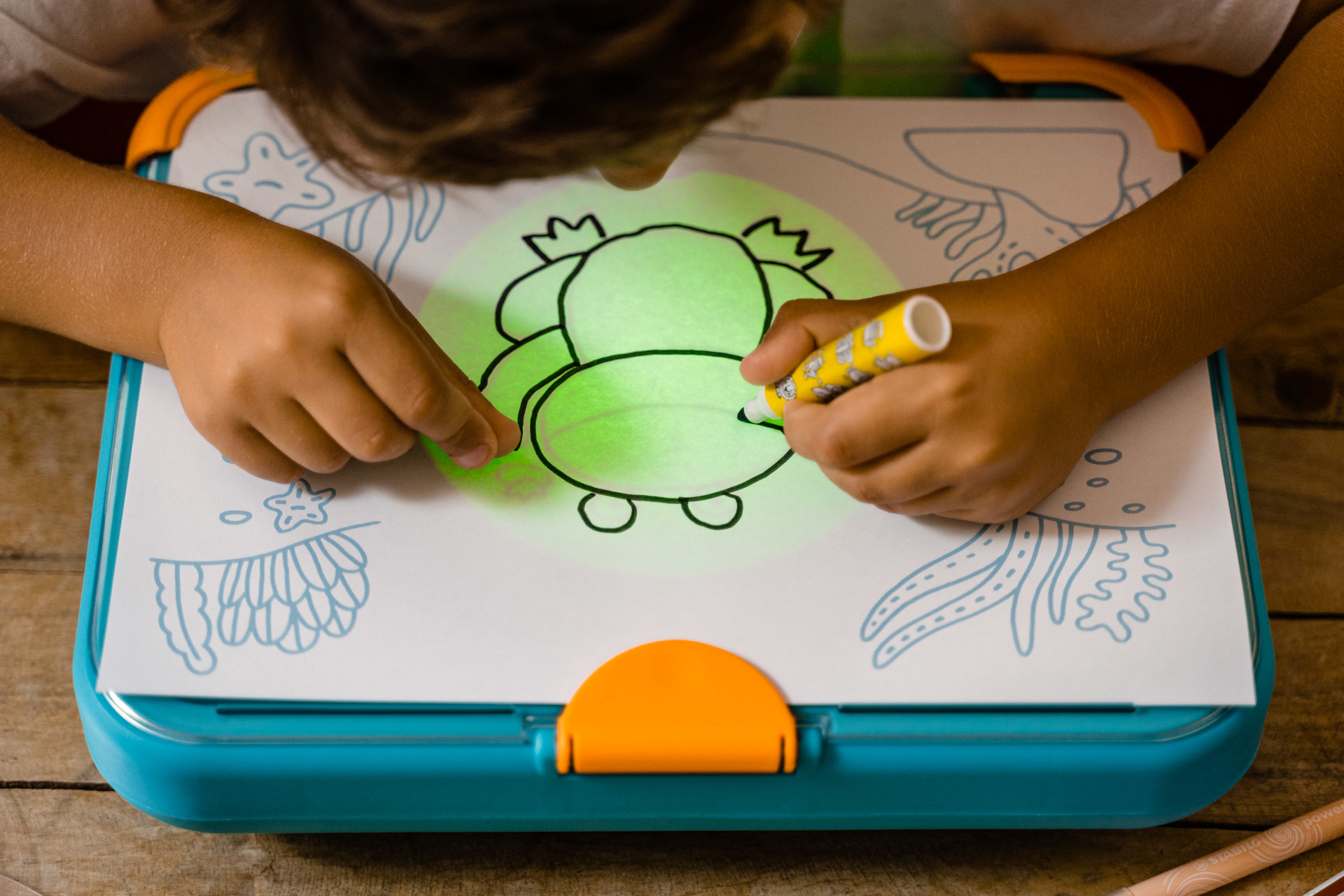 Dziecko rysuje żabę na podświetlanej tablicy do rysowania żółtym pisakiem. Tablica jest niebieska.