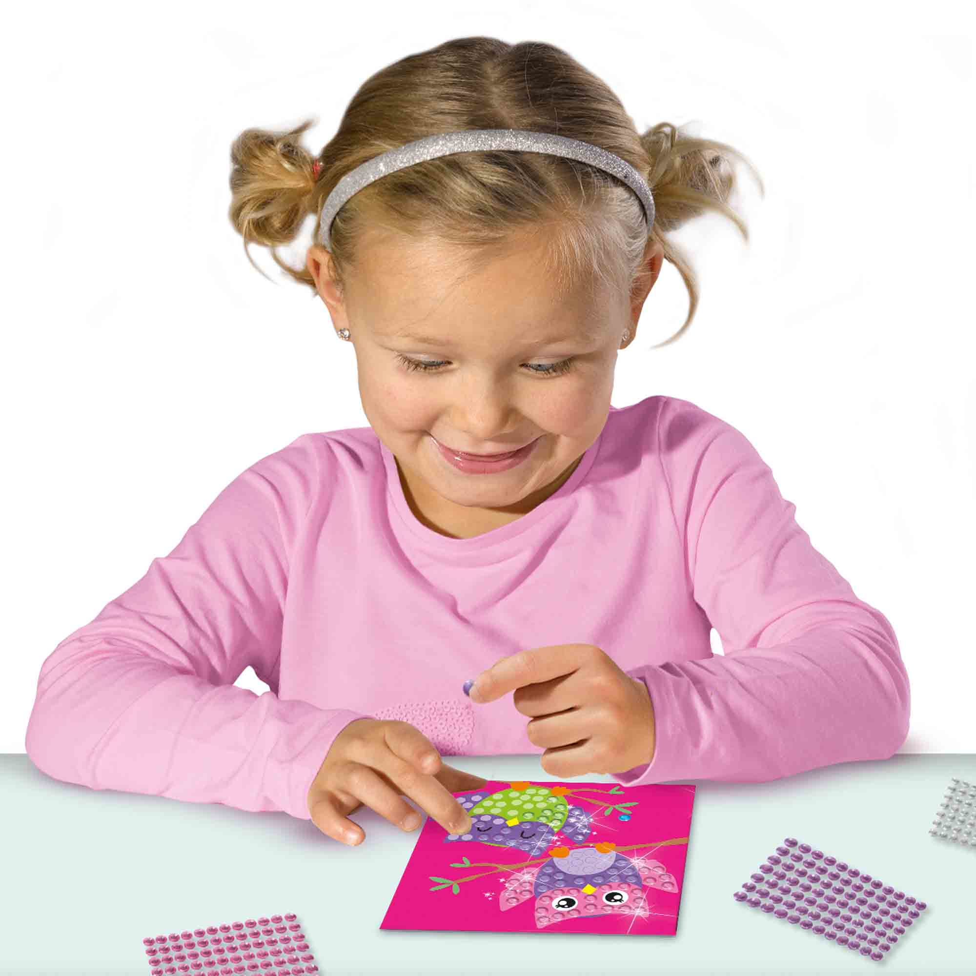 Dziewczyna maluje diamentami. Dodaje klejnoty do obrazu sowy. Na stole są inne obrazy i klejnoty.