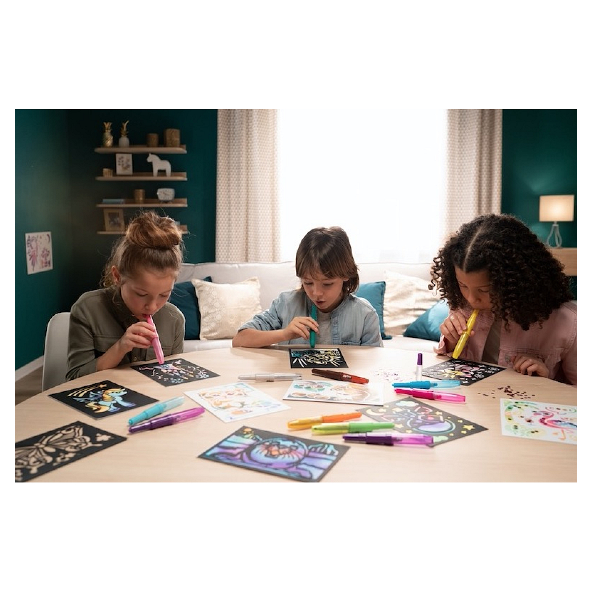 Trójka dzieci przy stole używa markerów do kolorowania na czarnych kartkach. Widoczne są różne przybory artystyczne.