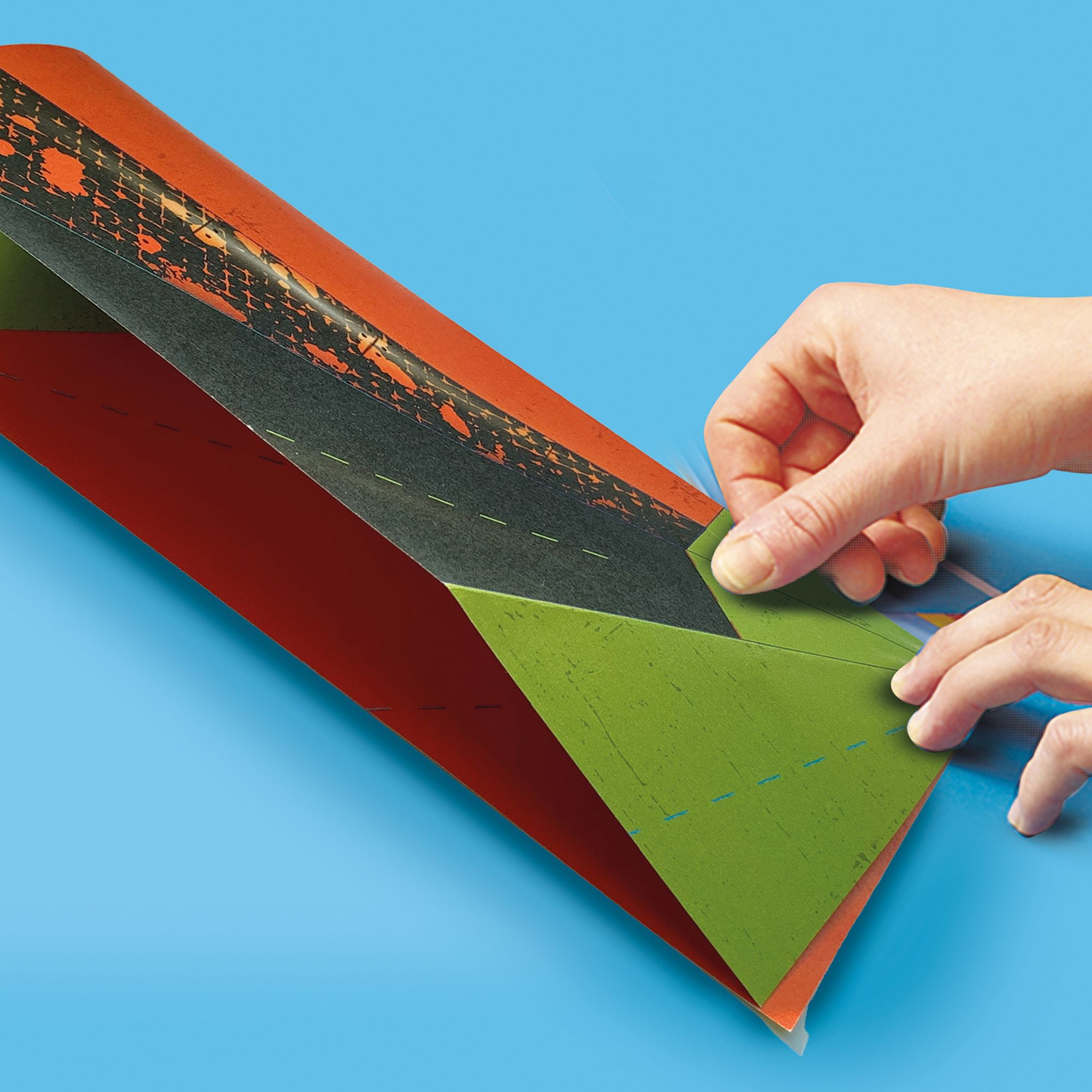 Dłonie składające papierowy samolot z pomarańczowym, zielonym i czarnym wzorem na niebieskim tle.