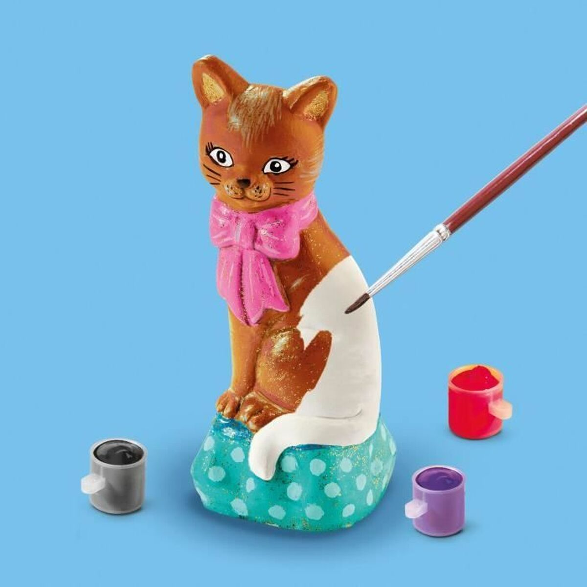 Una statuina di gatto viene dipinta con un pennello. Contiene barattoli di vernice e una statuina di gatto seduta su una superficie blu.