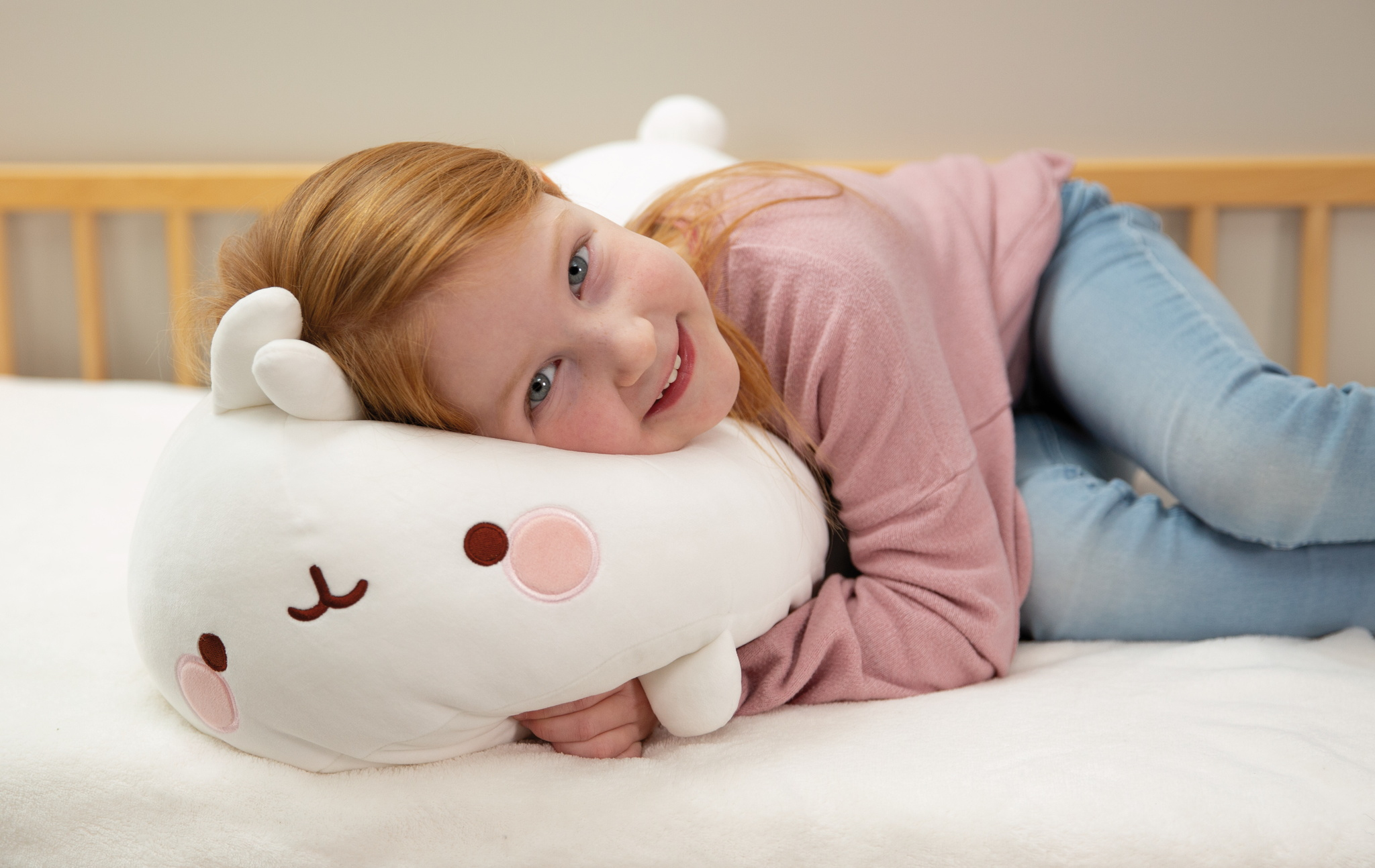 Dziewczyna leżąca na łóżku, uśmiechnięta i trzymająca białą, pluszową poduszkę w kształcie królika.