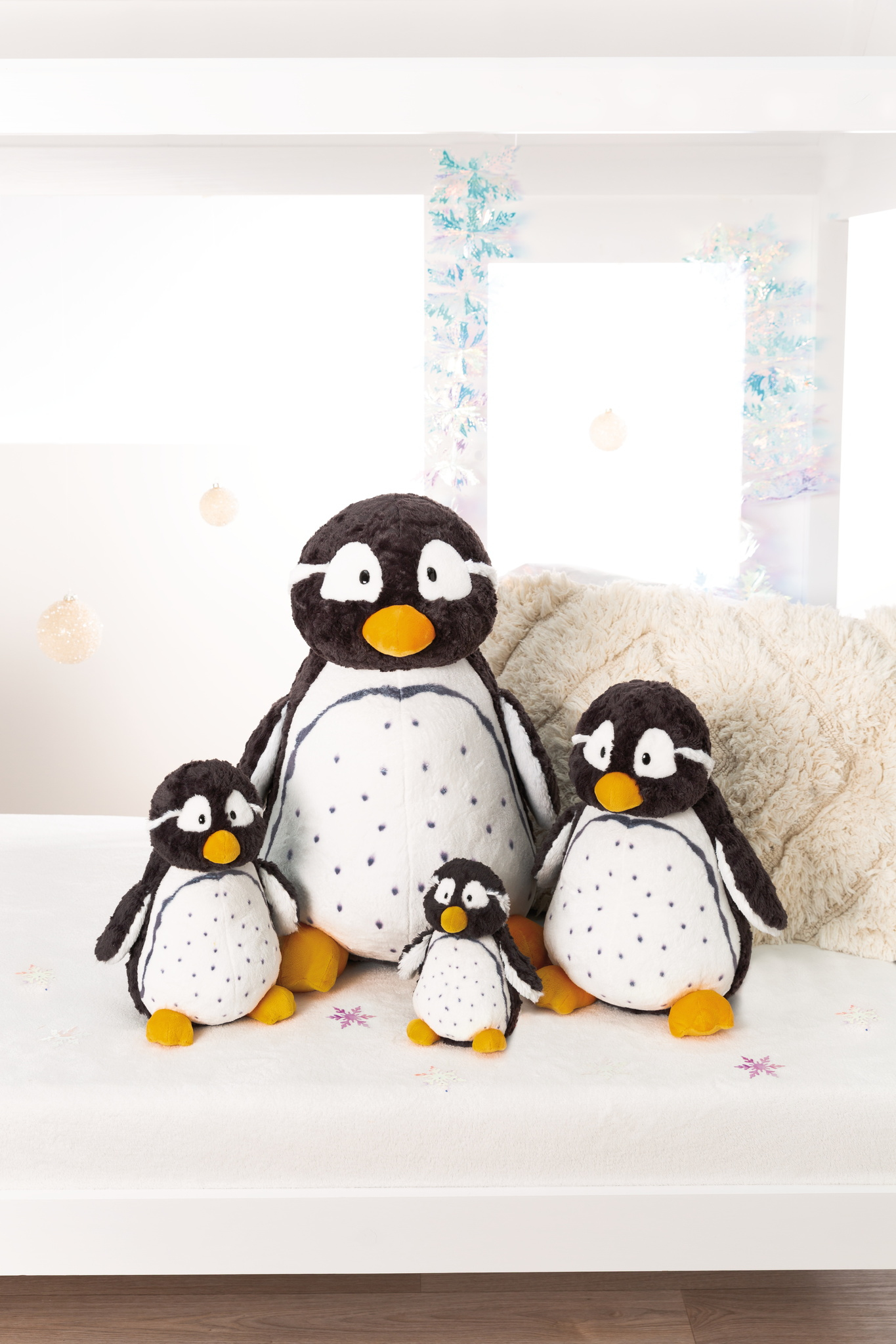 Cztery pluszowe zabawki pingwinów w różnych rozmiarach. Są czarno-białe z pomarańczowymi stopami. Białe tło.