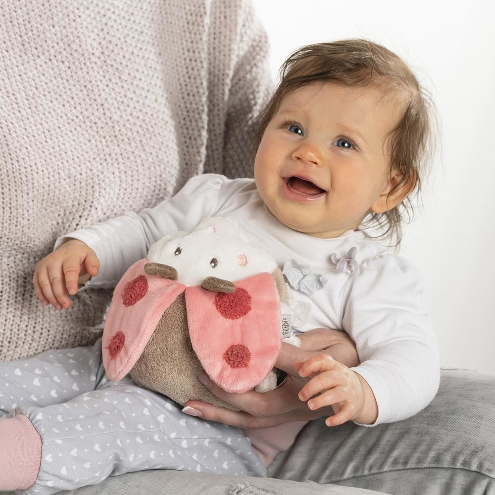 Niemowlę uśmiecha się, trzymając zabawkę w kształcie biedronki. Biedronka jest różowa, brązowa i biała.
