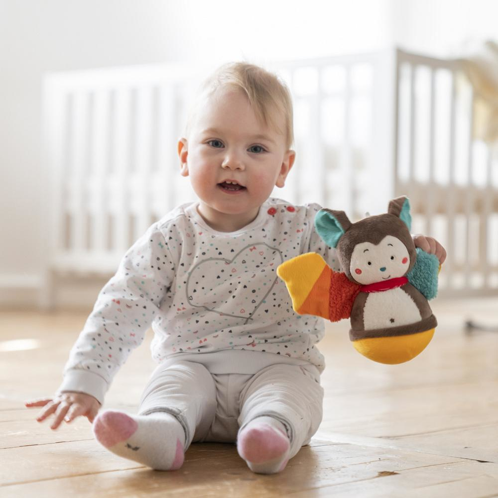 Niemowlę siedzące na podłodze, bawiące się zabawką w kształcie nietoperza. Niewyraźne tło.