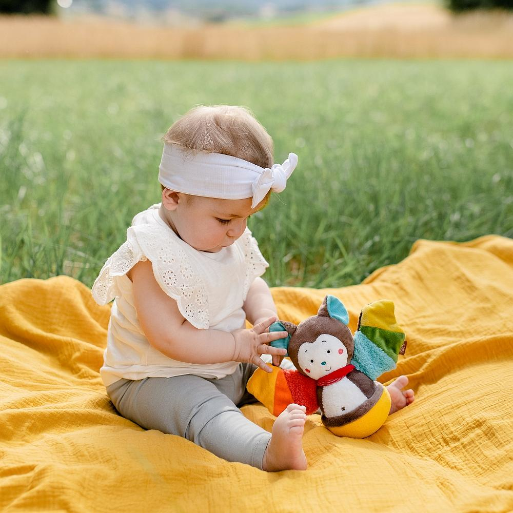Niemowlę siedzące na żółtym kocu, bawiące się zabawką w kształcie nietoperza, trawniaste tło.