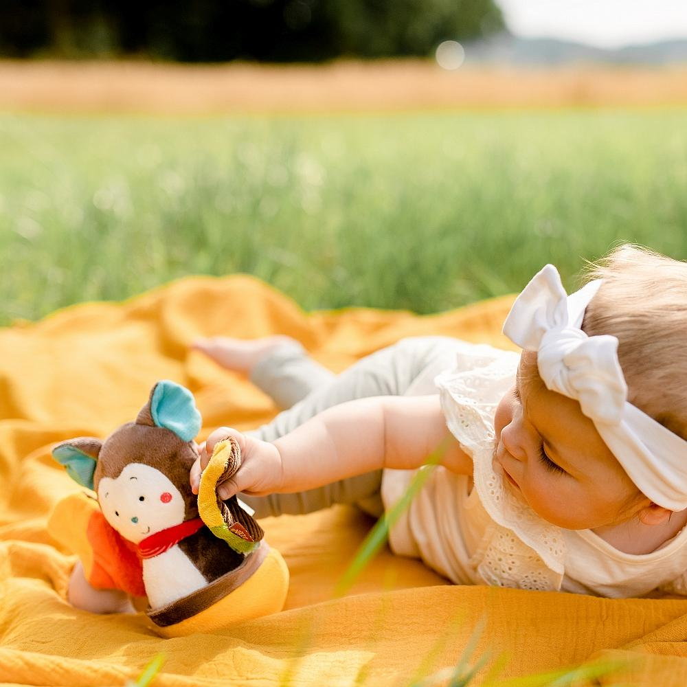 Dziecko na żółtym kocu z zabawką małpką. Zielona trawa w tle.