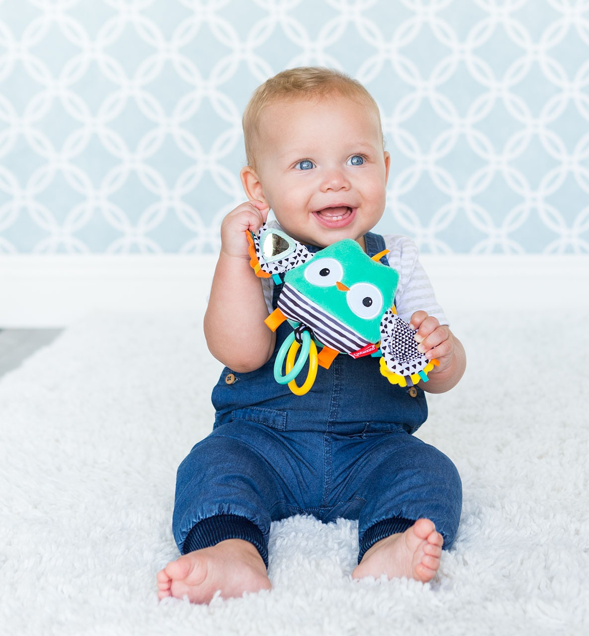Dziecko w dżinsowych ogrodniczkach trzymające zabawkę sowy, siedzące na białym dywanie, z niebieską ścianą.